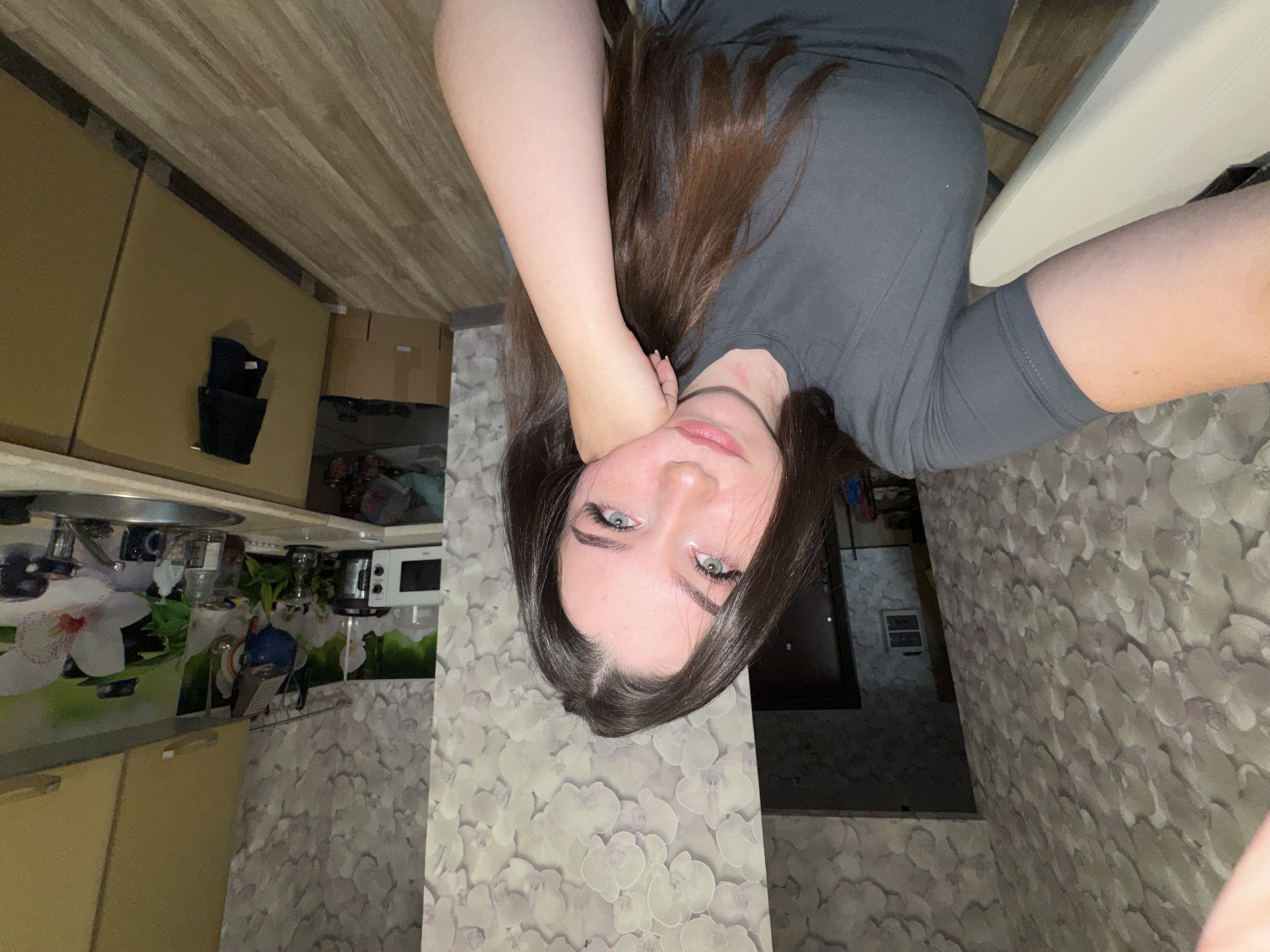 A young woman with long, dark brown hair and light blue eyes is taking a selfie in what appears to be a kitchen. She is wearing a dark grey t-shirt. Her right arm is resting on her cheek, and she is looking directly at the camera with a pensive expression. The background shows floral wallpaper, a kitchen counter with appliances and cabinets, and a wooden floor. The location appears to be an indoor domestic setting.
