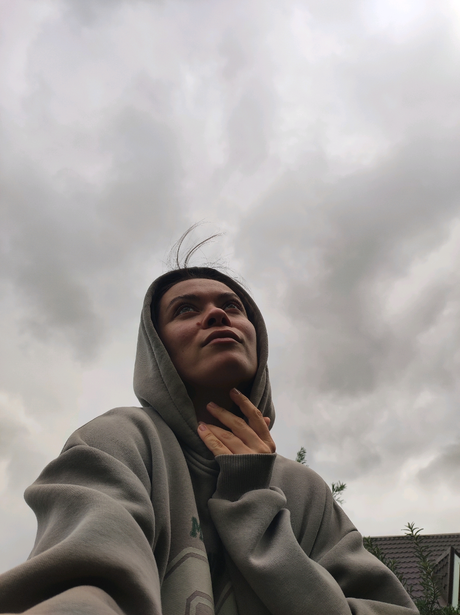 A young woman wearing a gray hooded sweatshirt looks up at a cloudy sky. Her left hand is raised to her neck. The image is taken from a low angle, with the sky occupying the majority of the frame. In the background, there is a glimpse of a house with a tiled roof and some greenery. The location is identified as Fokino, Russia, though no specific landmarks are visible to confirm this.