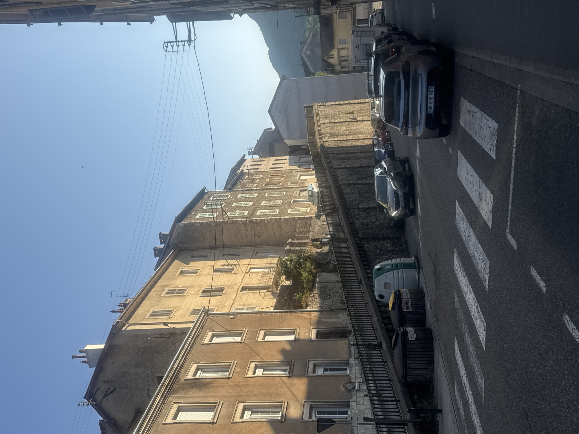 In Saint-Claude, France, a street scene unfolds under a clear blue sky with sunlight casting shadows. The foreground features a pedestrian crossing on asphalt, with white stripes marking the path. To the right of the crossing, a gray Peugeot car is parked. Further down the street, several other cars are lined up along the curb. On the left side of the street, a raised sidewalk or embankment is visible, bordered by a black metal railing. Two trash bins, one dark and ribbed and another green and white, sit on the embankment. Beyond the embankment, the facade of a multi-story building with numerous windows can be seen. The building appears to be made of light-colored stone and has a classic architectural style. In the background, more buildings and a hillside with trees are visible, suggesting a town nestled in a valley. Power lines stretch across the upper portion of the image. The overall impression is of a quiet, sun-drenched street in a French town.