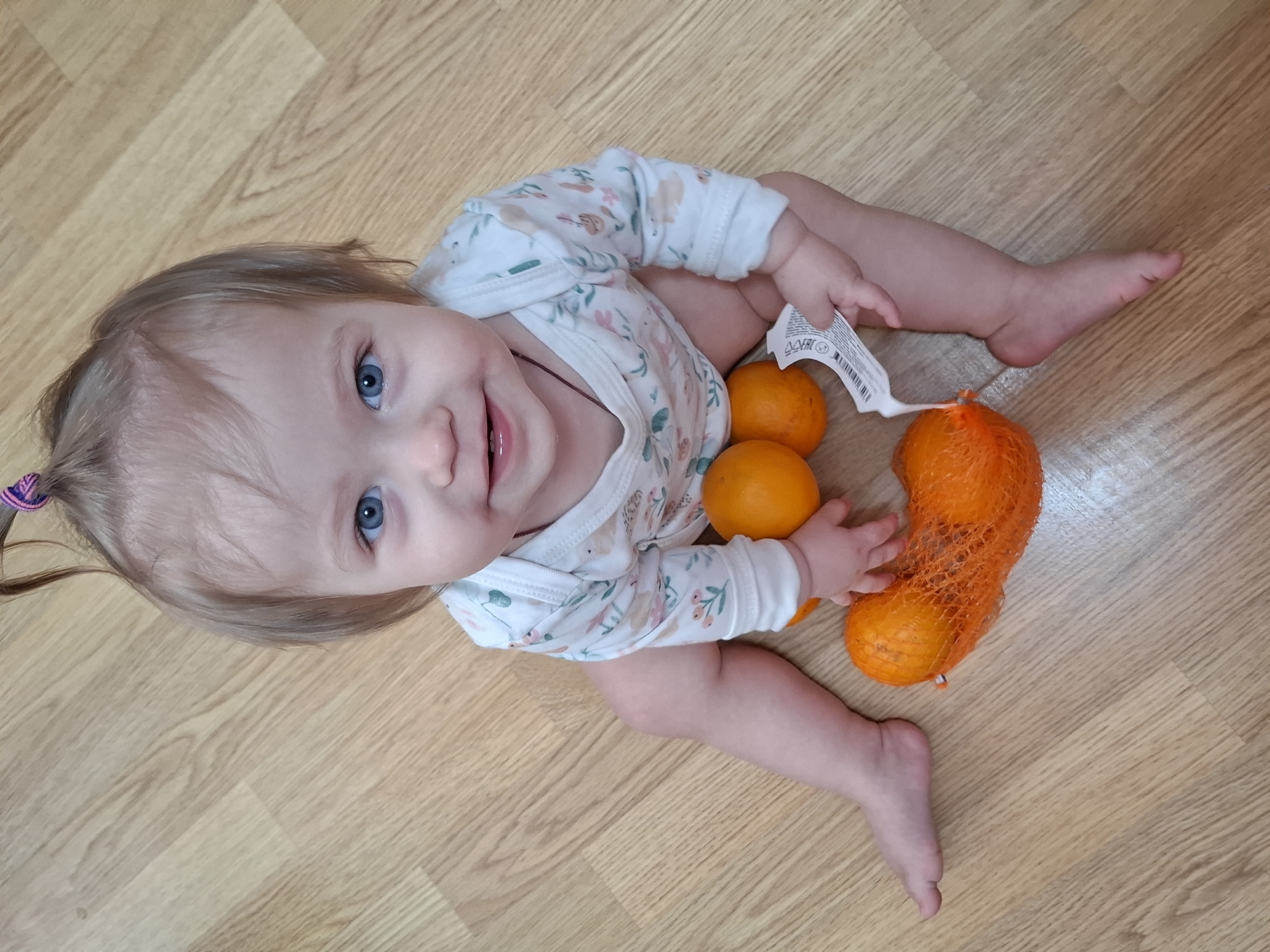 A baby with blue eyes and blonde hair sits on a wooden floor next to a bag of tangerines. The baby wears a white, floral-patterned onesie. The background is a wooden floor with a light brown color. The setting is unknown but possibly a residence. There is no verifiable indication of the location being Chaikovsky, Russia.