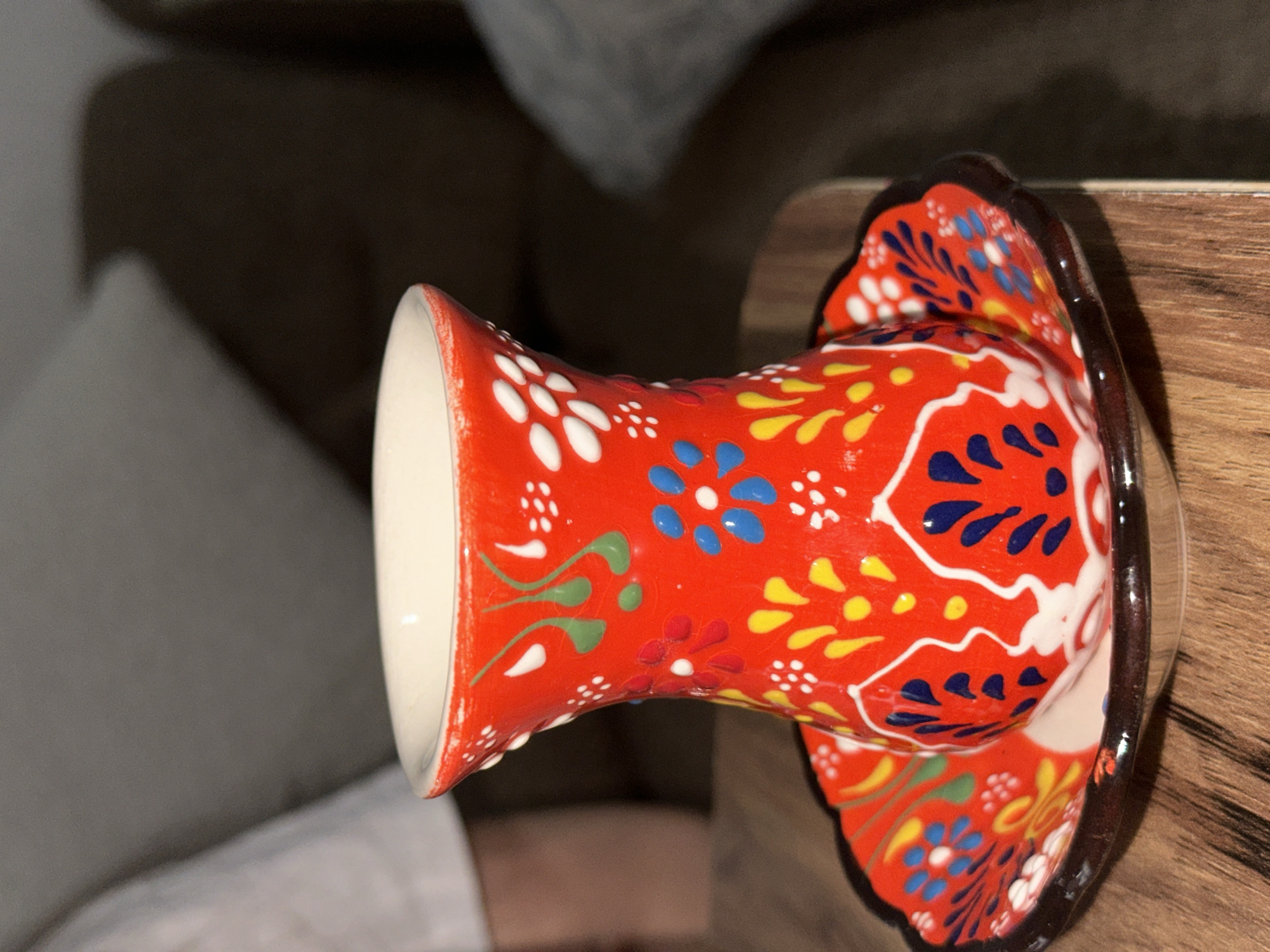 An eye-level, close-up shot focuses on a colorful ceramic vase, likely of Turkish origin, resting on a light brown wooden surface. The vase is predominantly red with a variety of floral patterns in blue, yellow, green, and white. It has a bulbous base that flares out, and a tall, slender neck that opens at the top. The rim is white. The vase appears to be resting on a circular base, with the same colorful floral patterns as the vase itself. The base has a dark brown rim. The wooden surface the vase is on appears to be a side table or shelf. The background is a blurred out image of an unidentifiable object.
There is no information that would allow this object to be placed in a specific location in Esenyurt, Turkey. The photograph is a close-up of the vase. The location could be anywhere.