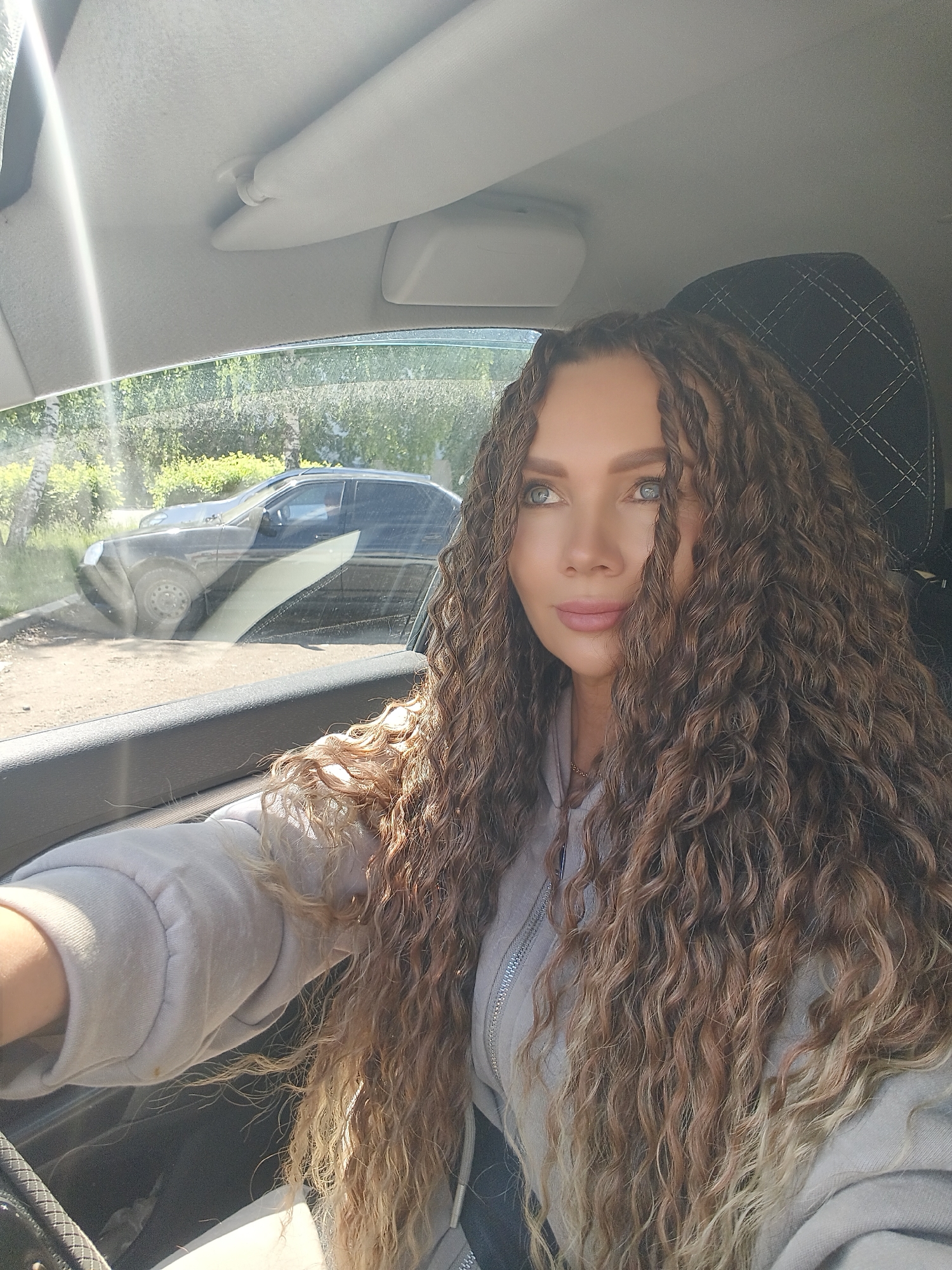 A woman with long, voluminous, curly hair, multi-toned from dark brown to lighter caramel and blonde towards the ends, is captured in a selfie-style shot from inside a car. Her light-colored eyes (blue/green) are directed forward and slightly to her right, giving an attentive or contemplative expression. She wears a light grey or beige hooded sweatshirt.
The setting is the interior of a car, with the woman positioned as if in the driver's seat, with her arm resting near the steering wheel. A sun visor and the car's headliner are visible above her. A black car seat headrest with white stitching is behind her. Bright sunlight streams in from the upper left, creating a lens flare on the windshield.
Through the car window on the left, an outdoor scene is visible. Another dark-colored sedan is parked on what appears to be an unpaved or dirt ground, backed by lush green trees and bushes, suggesting a daytime setting with good weather. The overall impression is a casual moment taken during a drive or while parked. There is no visible text in the image.