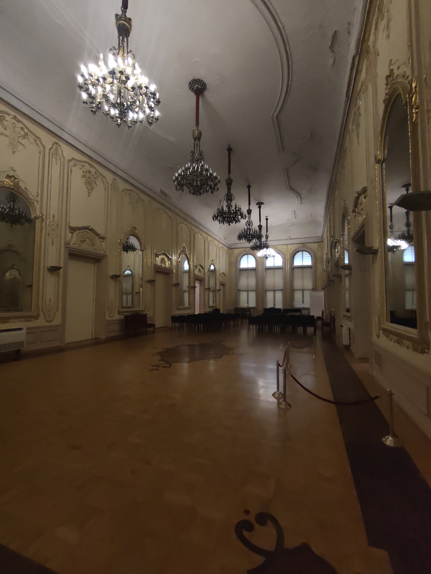 The image shows a grand, empty ballroom, likely within a historical building in Sofia, Bulgaria. The room features a high, ornate ceiling with several large, multi-tiered chandeliers, each illuminated. The walls are decorated with intricate gold molding and large mirrors, reflecting the room's opulence. Several arched windows with closed blinds line one side of the ballroom, admitting diffused light.
The floor is polished wood, showing some signs of wear and tear. In the foreground, a velvet rope barrier on stanchions separates the viewer's perspective from the main area of the ballroom.
Several rows of dark-colored chairs are arranged in the middle of the room, facing a blank wall, suggesting it's set up for a performance or presentation. Pianos are visible in the background, arranged in formation. Doors are interspersed along the walls, some of which are visible in the reflections of the mirrors. The overall atmosphere is one of stately grandeur and stillness, with no people present.