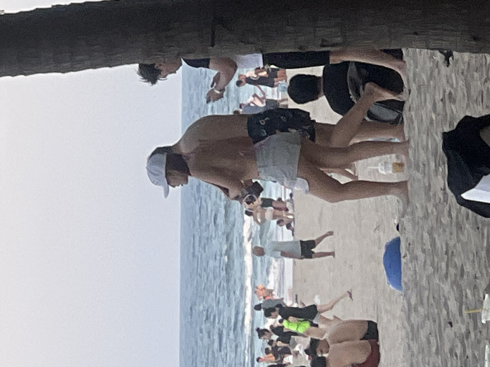 The image depicts a crowded beach scene in Đà Nẵng, Vietnam, with numerous individuals visible in the background wading in the ocean and walking along the shore. In the foreground, a couple is standing on the sand, with the woman wearing a white cap, a patterned bikini top, and light-colored shorts. The man beside her is shirtless, wearing dark patterned shorts. They appear to be interacting, possibly looking at a phone the man is holding. To the right, a portion of a tree trunk is visible, partially obscuring another person who is holding a phone and wearing a dark shirt with a white sleeve. The beach has sand, and the ocean is visible with waves. The sky is overcast and a light blue. Several other people are scattered across the beach, some sitting, some standing, and some in the water.