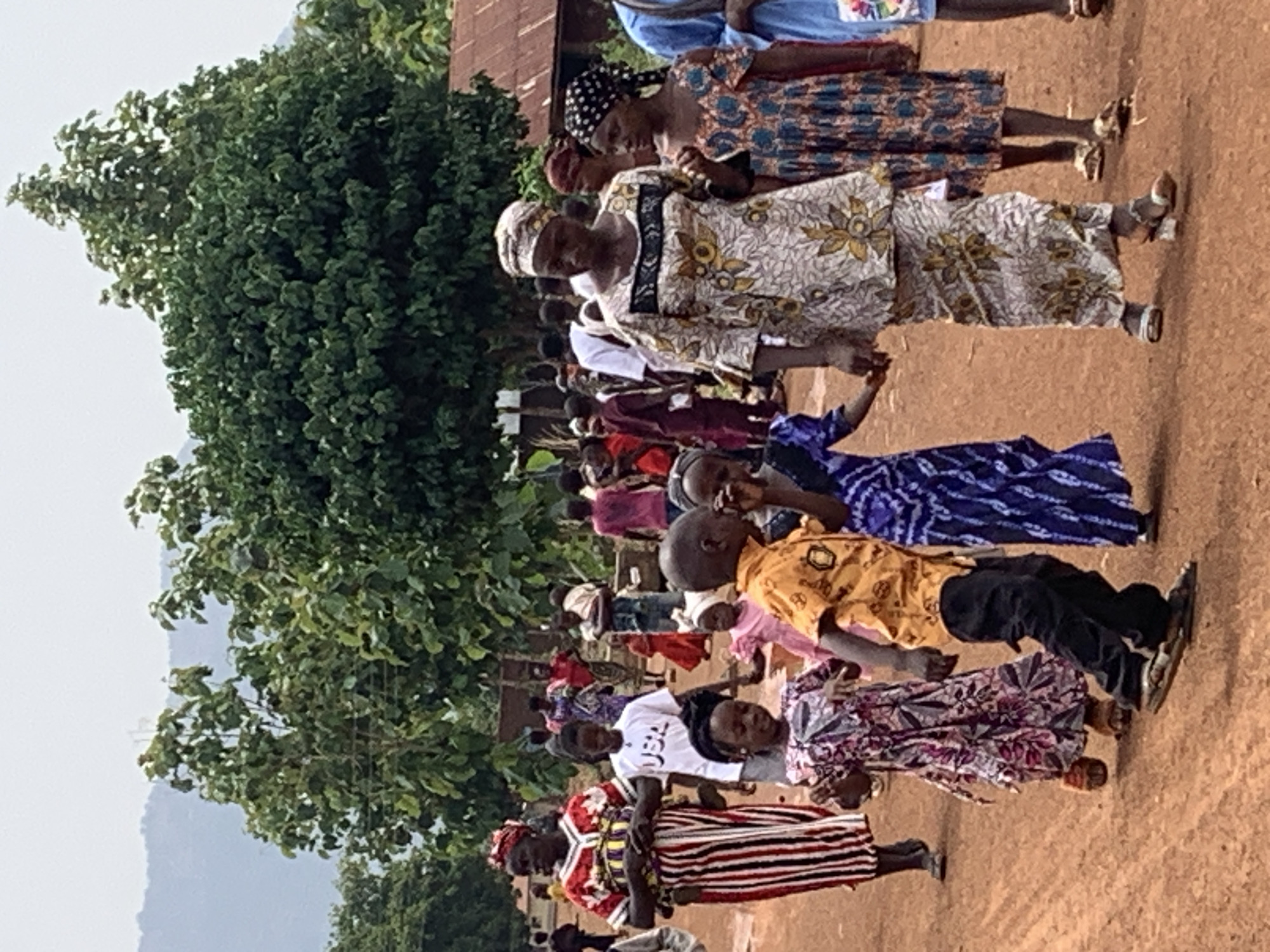 A group of people of various ages stand in a row on a dirt ground, presumably in Agban, Nigeria. A large green tree is visible on the left side of the frame. The people are wearing a variety of colorful clothing. Some are holding hands.