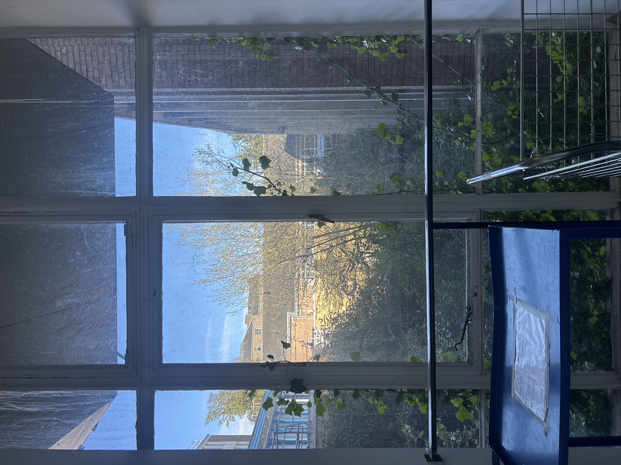 A photograph depicts a view through a multi-paned window, oriented vertically, in Dundee, United Kingdom. The window frame is white, and the panes are visibly grimy. Outside, a mix of brick buildings and vegetation is visible, including trees and climbing plants on the window. A blue structure with a white sign attached is partially visible on the right side. A black pole runs vertically on the right.