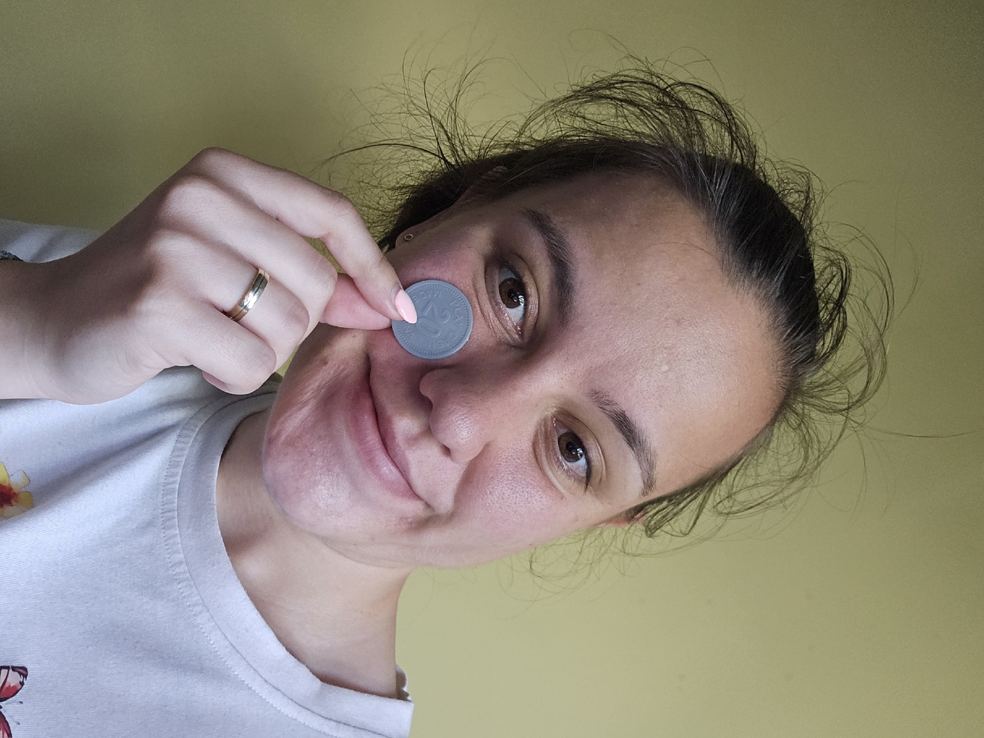 A young woman, with messy dark hair pulled back, is holding a grey coin to her left cheek with her right hand. She is wearing a white t-shirt with a butterfly design. Her left hand is resting on her chin and she is wearing a ring on her left ring finger. The background is a plain, light green wall. The image appears to be a selfie taken indoors. There is no identifiable information about the location in Sofia, Bulgaria.