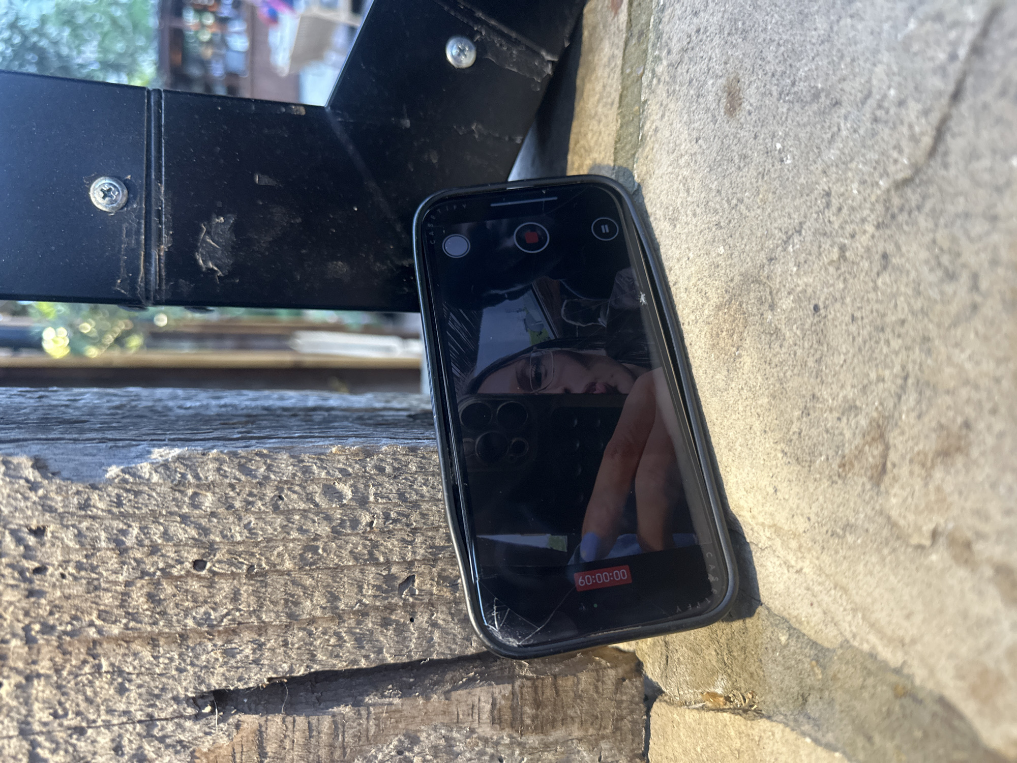 A damaged smartphone with a cracked screen rests between a rough wooden surface and a stone wall. The phone displays a paused video recording, reflecting a person and their hand with blue nail polish. The recording timer shows "60:00:00". A black metal structure with visible screws is positioned behind the phone, casting a shadow on the wooden surface. The location is unknown.