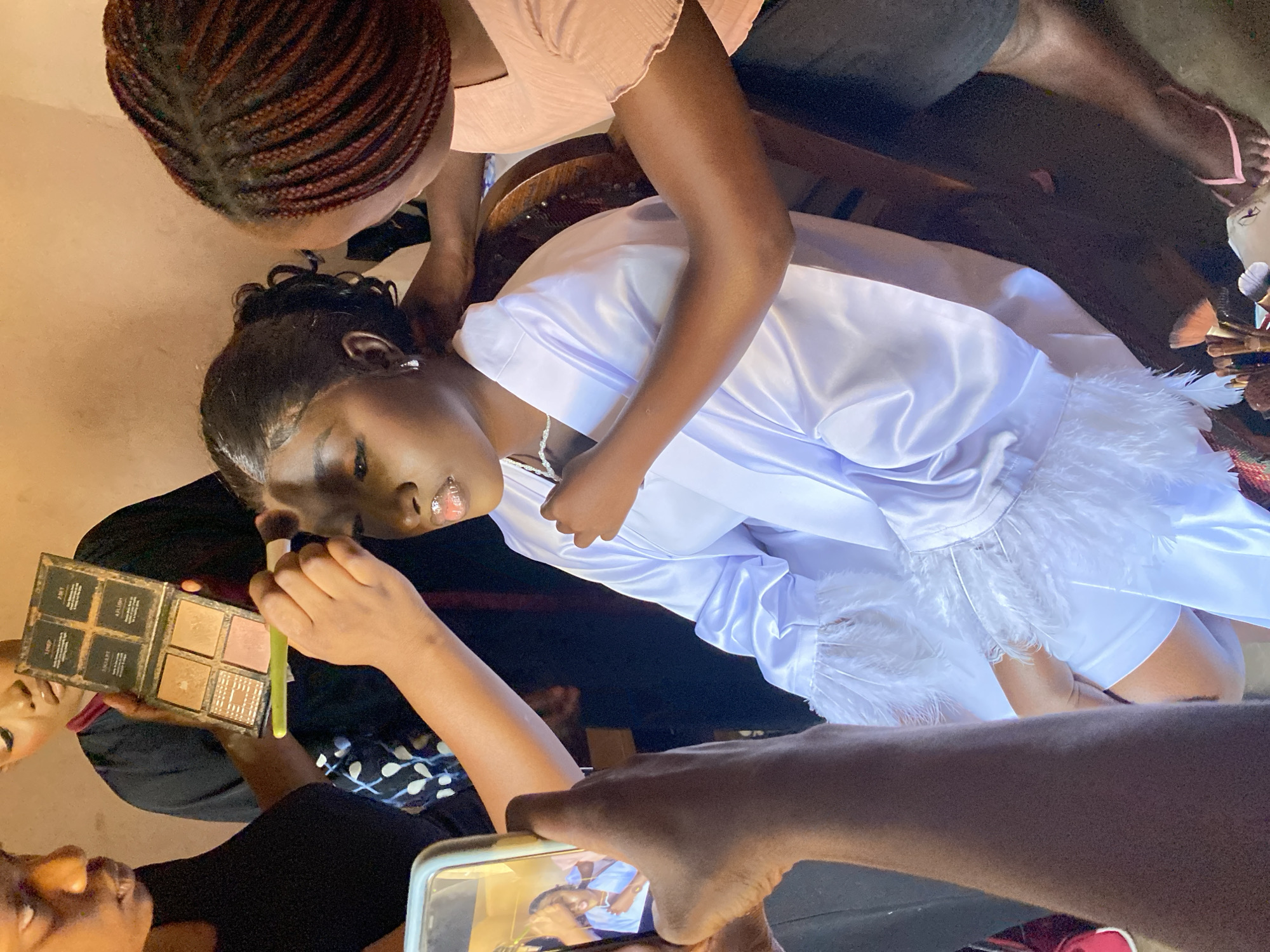 The scene appears to be a makeup session in progress, potentially in Kuru Karama, Nigeria. An individual lies prone, presumably receiving makeup. Two people attend to her: one applying makeup with a brush and a palette, and another adjusting her hair. A bystander records the scene with a smartphone. The person receiving makeup wears a white garment with feathered trim and a delicate necklace. The environment consists of neutral-toned walls and what appears to be a wooden chair.