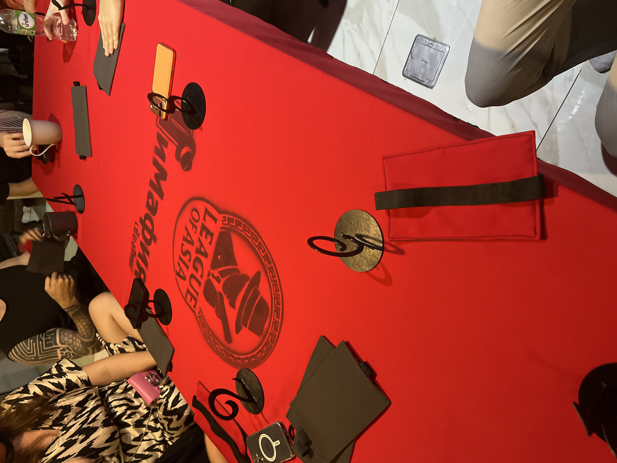 The image shows a group of people gathered around a bright red table with various items placed on it. The table appears to be set up for a game or activity, possibly a card game, given the presence of what looks like game mats and a deck of cards. The people are casually dressed, and their faces are not clearly visible, suggesting an informal gathering. One person is wearing a black sleeveless top with intricate tattoo designs on their arm. Another person is wearing a black and white patterned dress. There are drinks on the table, including a bottle of water and a mug, as well as a smartphone and some black holders. The background suggests an indoor setting, possibly a restaurant or a home, with tile flooring visible. The overall atmosphere is relaxed and social. The image does not provide any specific visual cues to pinpoint the exact location within Chiang Mai City Municipality, Thailand.