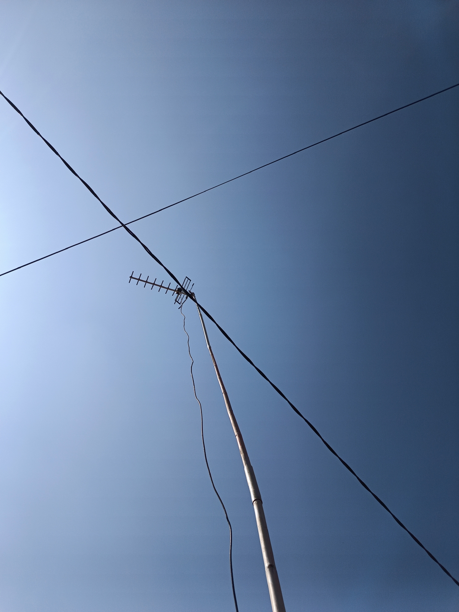 In Margajaya, Indonesia, a low-angle shot captures a clear, bright blue sky. Two thick, dark power lines stretch diagonally across the frame, one from the lower left to the upper right, and the other from the upper left to the lower right, forming an 'X' shape. Below and slightly to the right of the intersection of these power lines, a television antenna is mounted on a long, light-colored pole. The antenna itself is rectangular, composed of multiple horizontal bars. A dark cable hangs from the antenna, extending downwards along the pole. The pole appears to be made of bamboo or a similar segmented material. The scene is devoid of people.