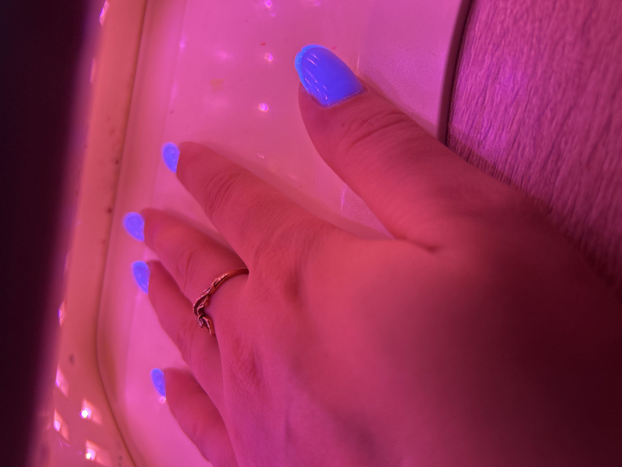A close-up, indoor shot under intense pink/red lighting captures a hand with bright blue polished nails. The hand is placed on what appears to be a white or light-colored surface, possibly a table or a device used for curing gel nail polish. A gold ring is visible on one of the fingers. The overall environment is dominated by the strong pink/red lighting, which obscures the details of the background and other objects. There is no verifiable information about the city of Chaikovsky, Russia, from the given image.