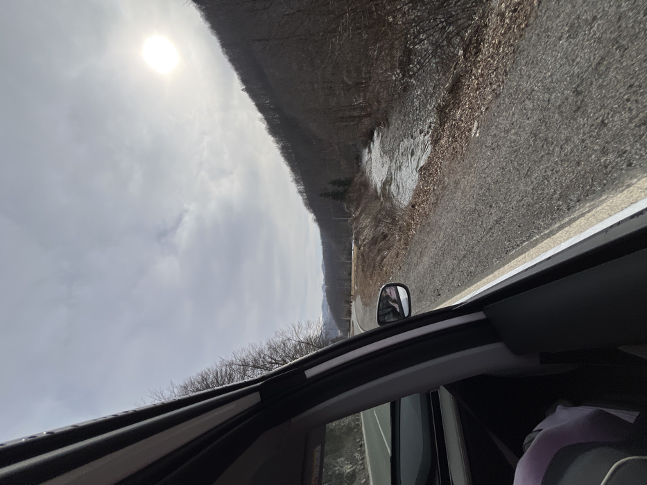 The image provides a view from the passenger seat of a vehicle in motion through a mountainous region in Lugovi, Montenegro. The car's black A-pillar and part of the passenger door frame are visible on the left. Through the windows, a two-lane paved road with a solid white fog line and a solid yellow center line is visible, curving to the left in the distance. To the immediate right of the road is an unpaved shoulder composed of gravel and brown, sparse vegetation. Further right, a flowing river or stream with a rocky bed runs parallel to the road; its banks display some brown foliage and potential ice patches. The landscape is dominated by steep hillsides and mountains covered predominantly with barren deciduous trees, indicating a cold season, possibly winter or early spring. A few coniferous trees are interspersed near the river. The sky above is partially cloudy, with a bright, intense sun visible in the upper right quadrant of the frame. A blurred reflection in the car's side mirror shows an indeterminate light-colored object or figure.
