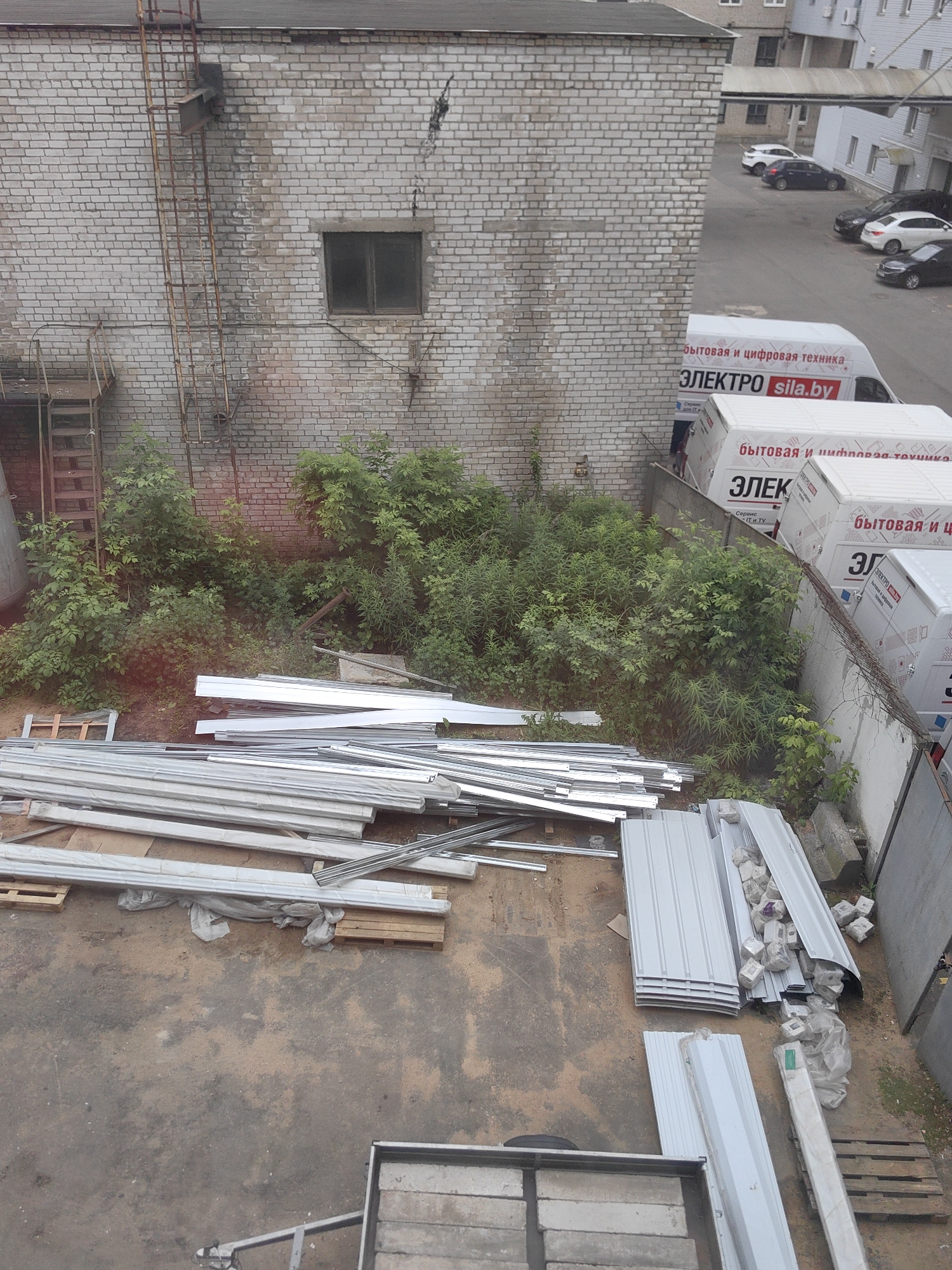 This is an aerial, outdoor, daytime photograph taken from a high vantage point, looking down at a construction or industrial area in Minsk, Belarus. In the background, a multi-story brick building is visible with a rusted fire escape ladder attached to its facade. A window is present on the building's wall. Beyond the building, several cars are parked on an asphalt surface. In the mid-ground, there are white delivery vans with Russian text, including "бытовая и цифровая техника" and "ЭЛЕКТРО sila.by". The foreground and mid-ground are dominated by piles of building materials, including long, slender metal sheets, corrugated metal panels, and wooden pallets. Lush green vegetation, including bushes and trees, grows in patches amongst the materials and along a retaining wall. The ground surface appears to be a mix of dirt and concrete.