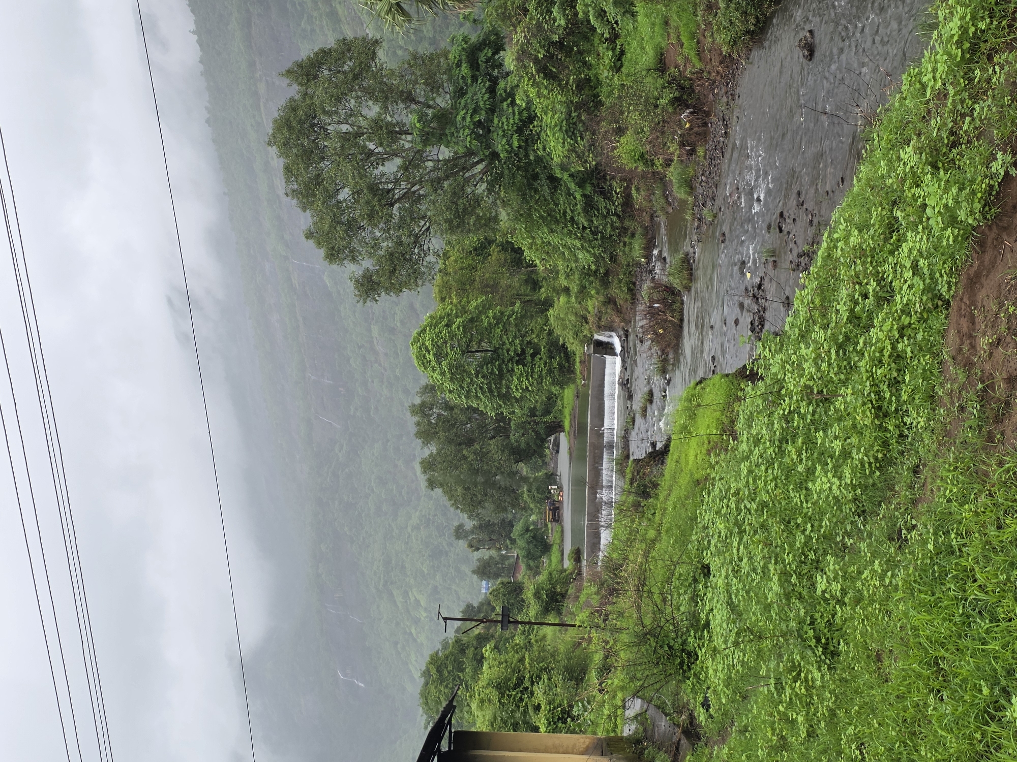 The image depicts a cloudy, overcast day in the rural outskirts of Sandshi, India. A small, concrete dam with water flowing over it bisects a rocky stream in the foreground. Lush green vegetation blankets the landscape, with dense trees and shrubs lining the banks of the stream. In the background, mist-shrouded hills rise, with faint waterfalls visible cascading down their slopes. To the left, a portion of a yellow-colored building with a dark roof is visible, possibly a small dwelling or roadside structure. A dirt path is discernible in the lower portion of the image, leading towards the stream. Overhead, power lines stretch across the frame, indicating a rural, yet connected, area. A yellow vehicle and a few people are visible near the dam in the middle ground, suggesting minimal human activity in this natural setting. The overall impression is one of serene natural beauty, characteristic of the monsoon season in a hilly region.