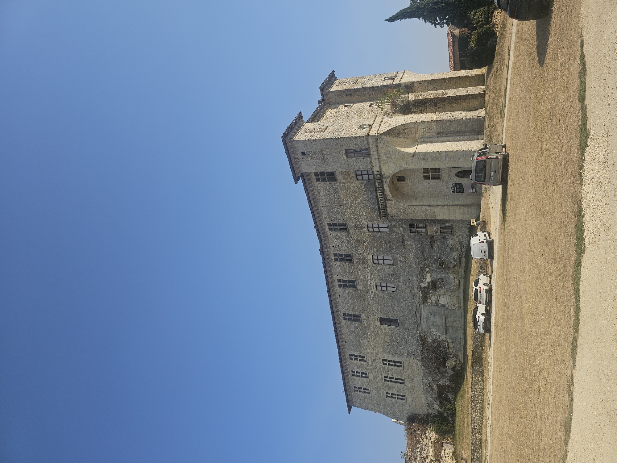 This is a full, eye-level, outdoor, vertical shot of a historic stone castle under a clear blue sky. The castle, built with light-colored stone blocks, dominates the frame from the middle ground to the right. It features multiple stories, numerous rectangular windows with dark panes, and crenelated battlements along the roofline. An arched entryway is visible on the lower facade. In the foreground, dry, golden-brown grass covers the ground, with a dirt path winding through it. Several vehicles are parked on the grass and path in front of the castle, including two white cars, a white van, and a silver minivan. A dark SUV is partially visible on the far right. The environment appears dry and sunny, suggesting a warm climate. The castle is located in Lavardens, France.