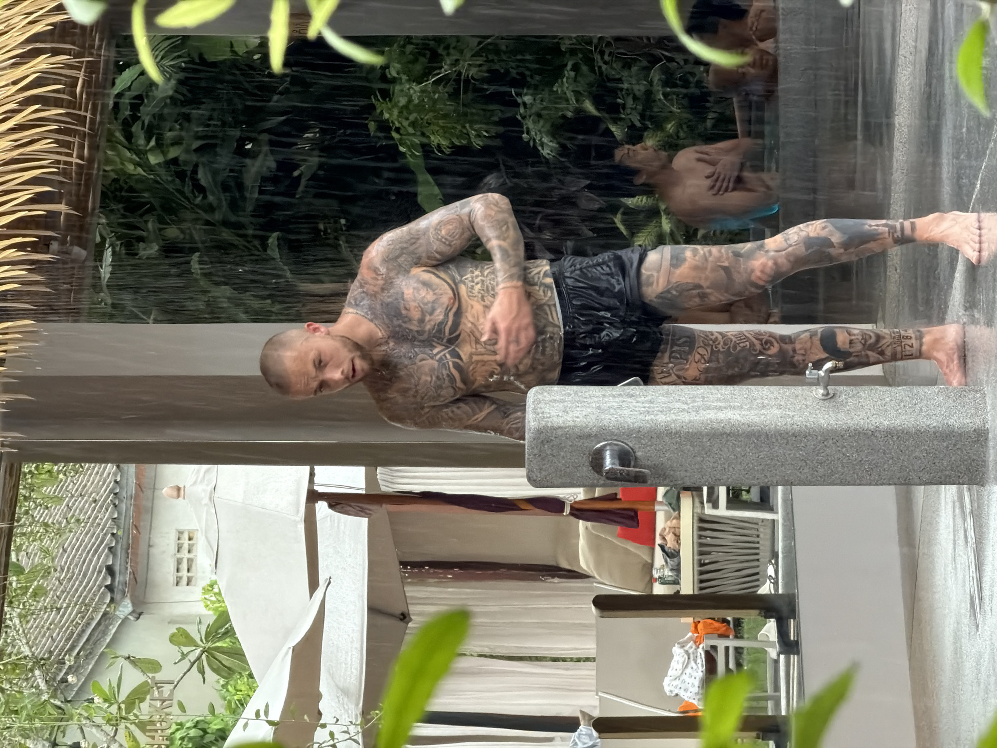 This media file shows two men in an outdoor shower, possibly in a tropical setting, given the lush greenery and what appears to be a tiled roof in the background. The primary subject is a heavily tattooed man, facing the camera and looking down as he showers. He is wearing black swim trunks and his body is covered in intricate black ink. His skin appears wet and he is in the process of washing himself.
In the background, reflected in the wet surface of the shower area, another man is visible. He is seated and appears to be looking away from the camera. He is wearing blue briefs. The water is falling heavily, suggesting it is either raining or the shower is in full use. The scene is dimly lit, but the surrounding foliage indicates it is daytime, likely overcast due to the rain or the shower's effect on the light.
The setting includes a stone structure which is likely the shower base or a retaining wall, with a faucet visible on it. There are also hints of awnings or cabanas, with white fabric draped around, suggesting a resort or private villa. Further in the background, there are glimpses of buildings and more vegetation. The overall mood is relaxed and personal, focused on the act of showering in a natural, outdoor environment. No visible text is present in the media file.