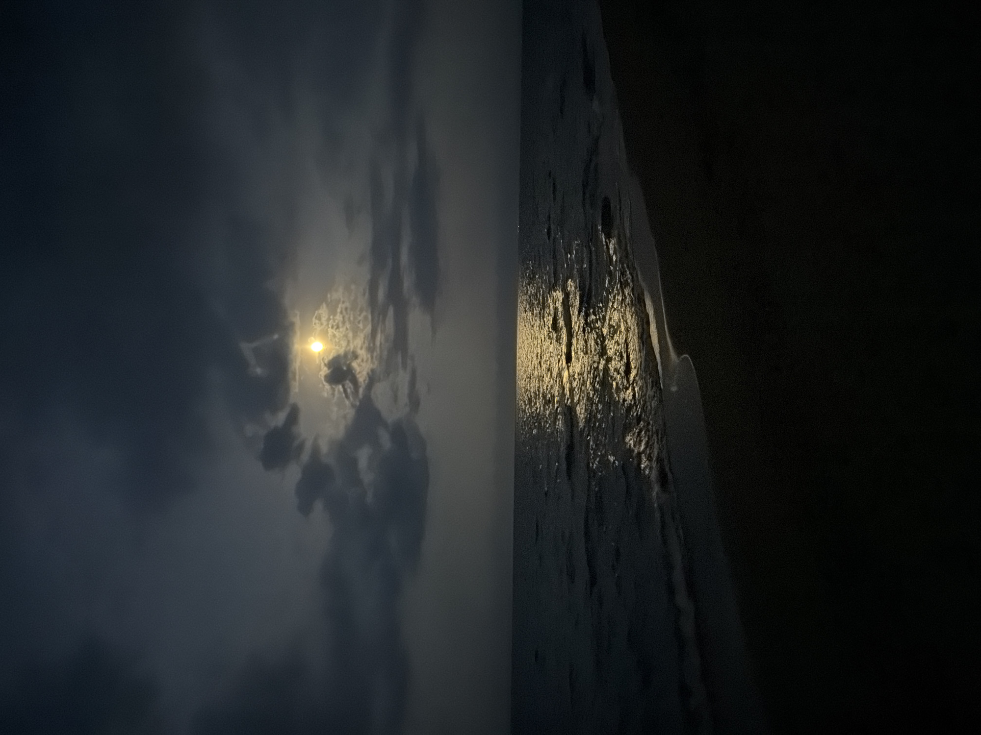 The image depicts a tranquil night scene at a beach. The moon, partially obscured by dark clouds, casts a soft, golden glow onto the gentle waves of the ocean. The water reflects the moonlight, creating a shimmering path across the surface that leads towards the sandy shore. The beach itself is dark and silhouetted, with the faint outline of the land meeting the sea. The overall atmosphere is serene and mysterious, evoking a sense of peace and solitude. The location is described as Unknown, Unknown.