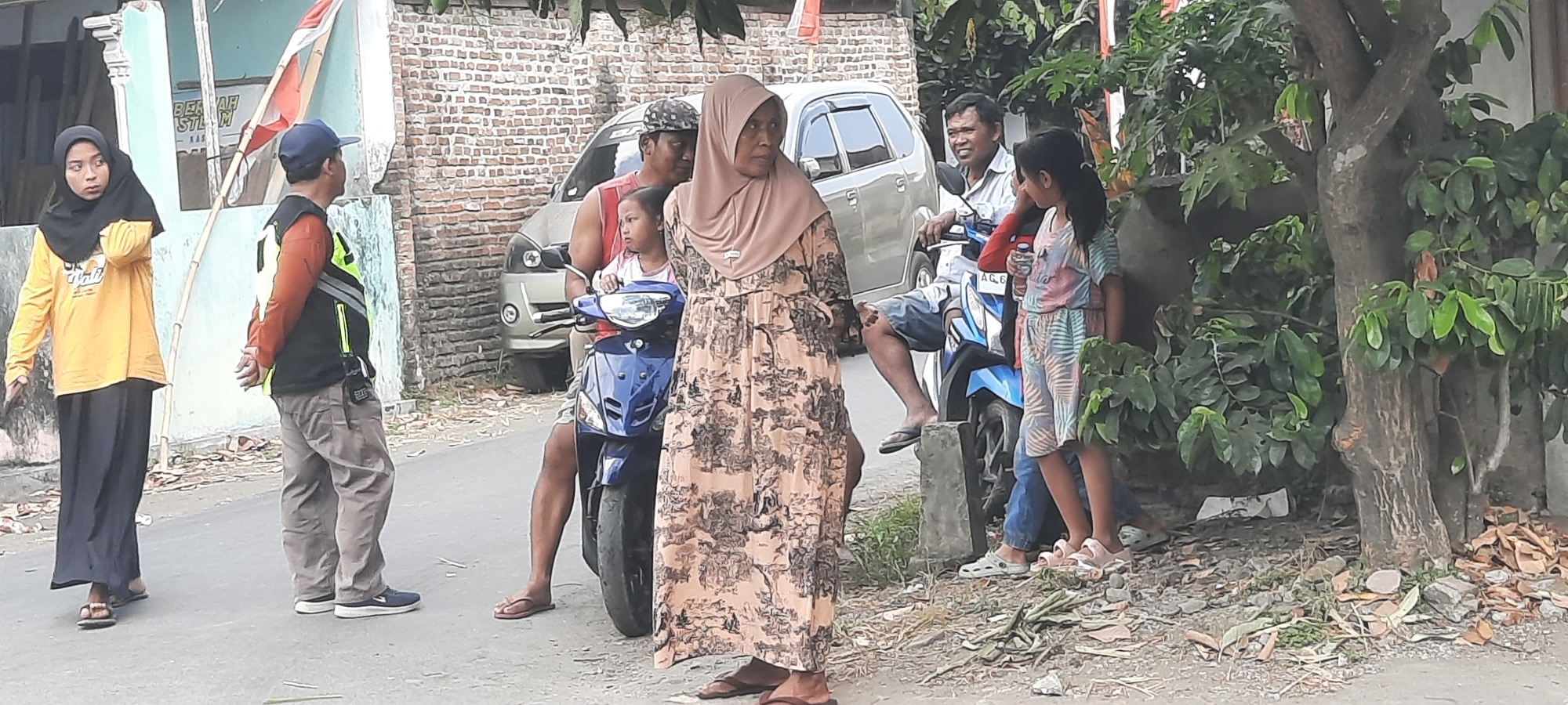 In the city of Jatitengah, Indonesia, a scene unfolds on a street lined with brick buildings and foliage. A group of people are gathered near a road. On the left, a woman wearing a black hijab and a yellow long-sleeved shirt stands with her hand holding a dark object. Next to her, a man in a red and black safety vest and a blue cap looks to his right. In the center of the image, a blue motorcycle is parked. A child is seated on the motorcycle, held by a man wearing a red sleeveless shirt and a patterned cap. To the right of the motorcycle, a woman in a beige floral dress and hijab is visible. Further right, a young girl in a colorful dress stands looking towards the motorcycle. Behind the people and motorcycle, a silver car is parked. The ground is paved and scattered with debris, including leaves and broken bricks. A large tree with dense green leaves is situated to the right of the scene, providing shade.