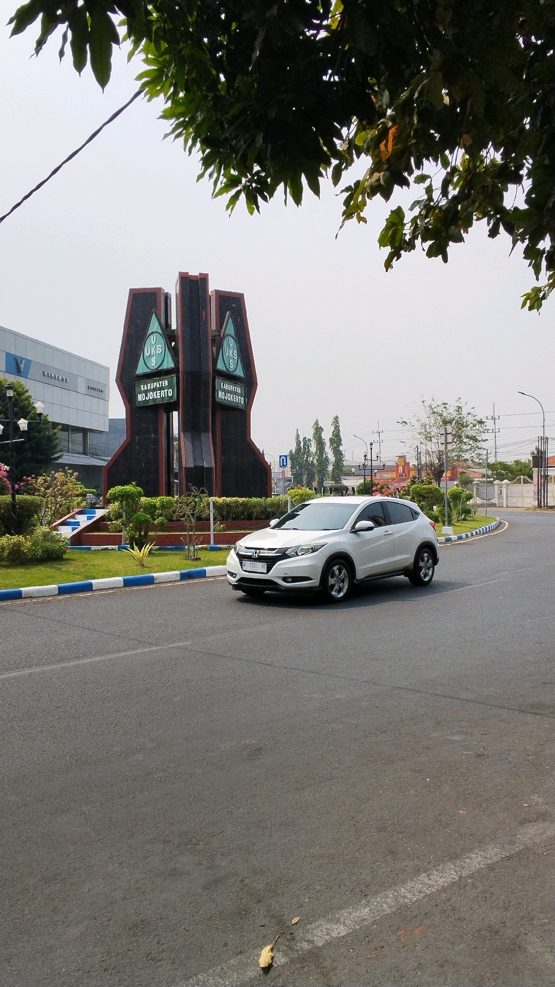 A white Honda HR-V drives down an asphalt road in front of the Mojokerto Regency monument. The monument is dark brown and black with green triangular panels bearing the letters "U K S" and the words "KABUPATEN MOJOKERTO". The vehicle is in motion, its front passenger side tire visible. In the background, there is a building with a "V" logo and the word "NISSAN" on its facade, as well as trees, streetlights, and a clear sky. The scene is outdoors, under daylight.