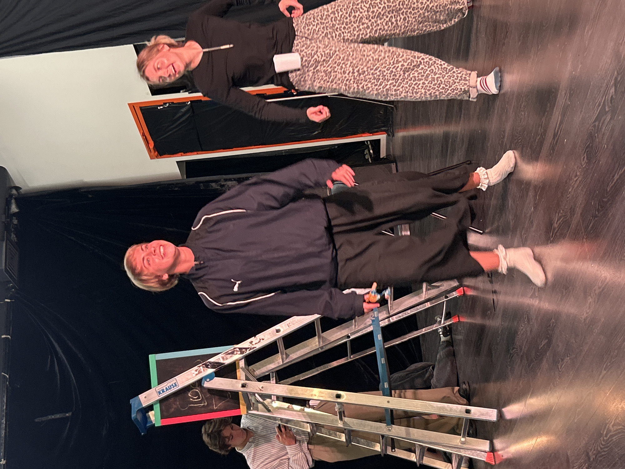 Three individuals are on a wooden stage. In the center foreground, a young person with blond hair and a playful expression is wearing a dark blue athletic jacket with white trim, dark pants, and white socks. To the right, a young person with blonde hair tied back is wearing a black long-sleeved shirt with a zipper and leopard-print pants. To the left, a young person in a striped collared shirt and beige pants is looking at a phone while standing next to a step ladder. The ladder is positioned in front of a black backdrop and leans against a blackboard with drawings. The stage floor is dark and reflective, with dramatic lighting creating reflections. The overall environment suggests a theatrical or performance setting.