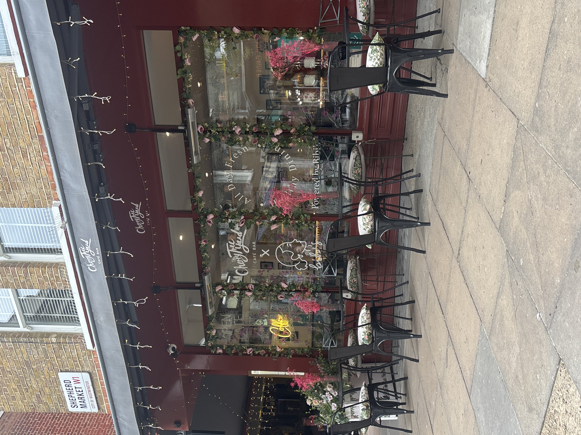 The image captures the exterior of "Crazy Tyyred" Cafe Bar, located on Shepherd Market W1 in the City of Westminster, London, United Kingdom. The cafe has a red façade with floral decorations around its windows. The storefront displays the cafe's name and logo, with "All Day Fresh Dining" visible in the window. Several black chairs and tables are arranged outside on the pavement, each chair having a floral cushion. A street sign indicating "SHEPHERD MARKET W1" is affixed to the brick building on the left side. Fairy lights are strung along the awning of the cafe.