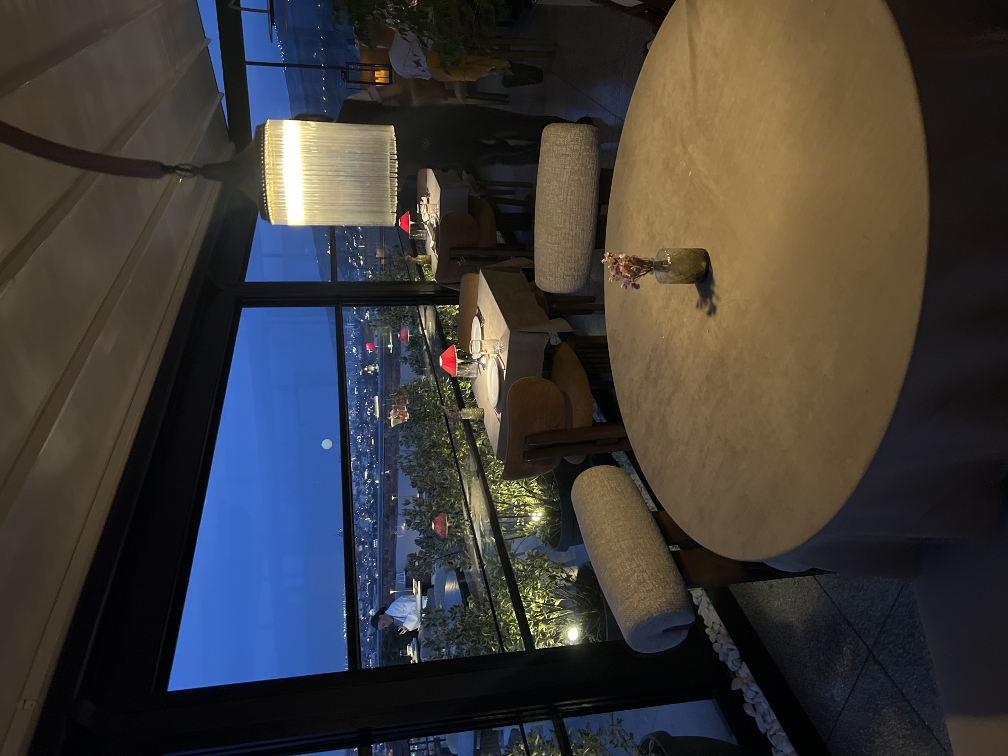 The image depicts an interior view of a rooftop restaurant in Tbilisi, Georgia, at dusk. The restaurant features a panoramic view of the city below, illuminated by the soft glow of twilight and scattered lights from buildings. Large windows offer an unobstructed vista of the urban landscape stretching towards the distant hills.
Inside, tables are set for dining. In the foreground, a round table is covered with a light-colored tablecloth, and a small glass vase with dried flowers serves as a centerpiece. Two upholstered chairs with light-colored fabric are positioned around this table.
In the mid-ground, another table is set with placemats, glasses, and what appear to be dinnerware, illuminated by a red-shaded lamp. A wooden-backed chair with a brown upholstered seat is visible. Further back, additional tables and chairs are arranged, suggesting a dining area. One table has a light-colored tablecloth and red lamps.
A person, appearing to be a server or patron, is visible in the background, partially obscured by the dim lighting and distance, wearing a dark outfit. Another person, a woman in a white robe or smock, is seated at a table near the window, looking down at her phone. She is part of the dining experience, engaged in her own activity.
The restaurant's ambiance is enhanced by decorative lighting. A large, cylindrical light fixture with a textured, ribbed shade hangs from the ceiling, casting a warm glow. Accent lighting highlights potted plants arranged along the balcony or terrace area. The outdoor space features a railing and what appear to be outdoor seating elements. The overall impression is one of an upscale dining establishment offering a scenic view of Tbilisi.