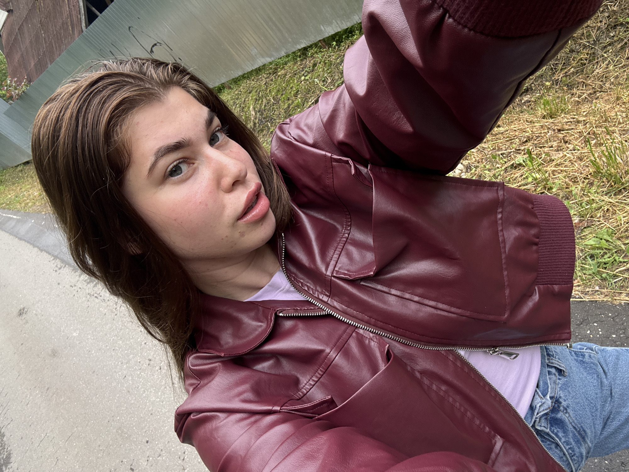 A young woman with long brown hair is captured from a high angle in what appears to be an outdoor setting. She is wearing a burgundy faux leather jacket over a light purple t-shirt and blue jeans. Her face is turned towards the camera, with her eyes looking directly at the viewer. The background shows a textured asphalt surface, green grass, and a corrugated metal fence. The overall impression is of a casual, everyday moment. Given the context of Мещёры, Russia, it's likely this scene takes place in a residential area or a park within the city.