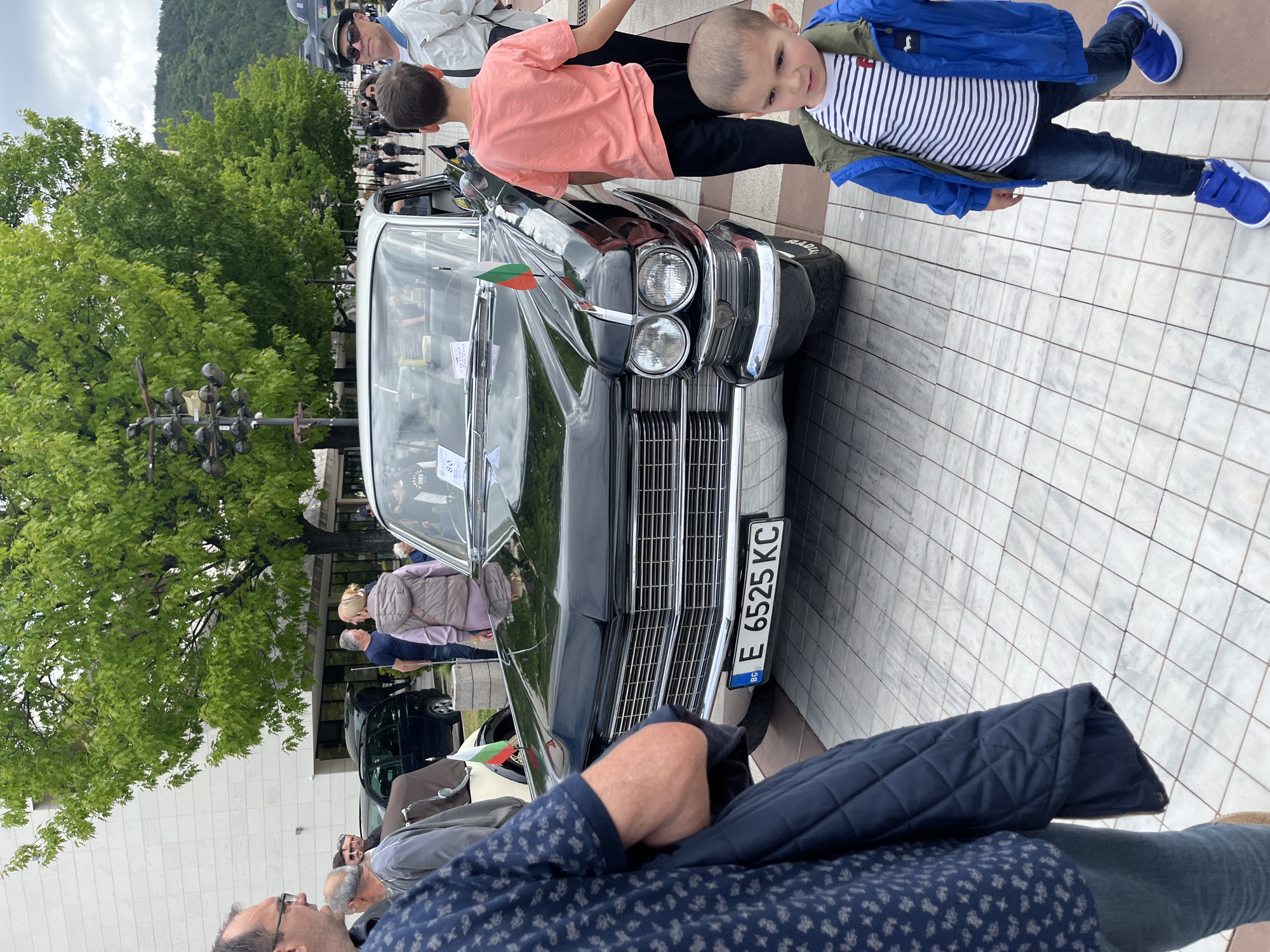 The image depicts a black vintage car, possibly at a car show or exhibition in a public urban space. A boy in a blue jacket and a striped shirt stands close to the car, with another child wearing a peach shirt leaning over the car's hood. People are milling around in the background. A license plate on the car reads "E 6525 KC BG". A tree and a lamp post are visible to the left, and a building façade is in the background. The location appears to be paved with light-colored tiles.
