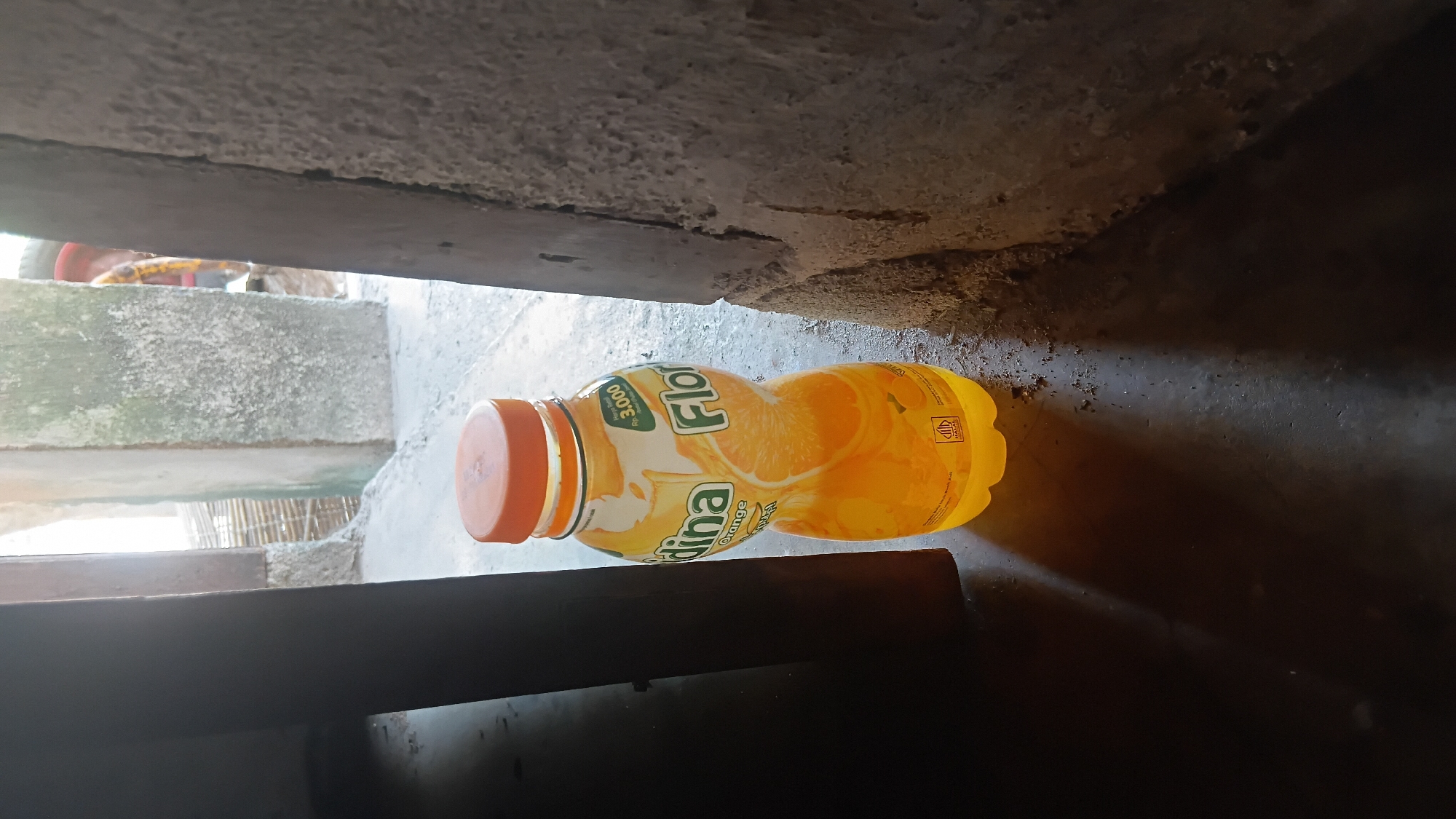 A single, partially filled bottle of orange juice, labeled "Floridina Orange", rests on a dark, textured surface, likely concrete or asphalt. The bottle has a bright orange cap and a label featuring images of orange slices and text. The environment appears to be an outdoor or semi-outdoor setting, with rough, grey concrete pillars and a dimly lit background suggesting a passageway or entrance. Light streams in from the right, casting shadows on the surface. The overall scene is static, with no discernible activity or individuals present. The location is identified as Prabumulih, Indonesia.