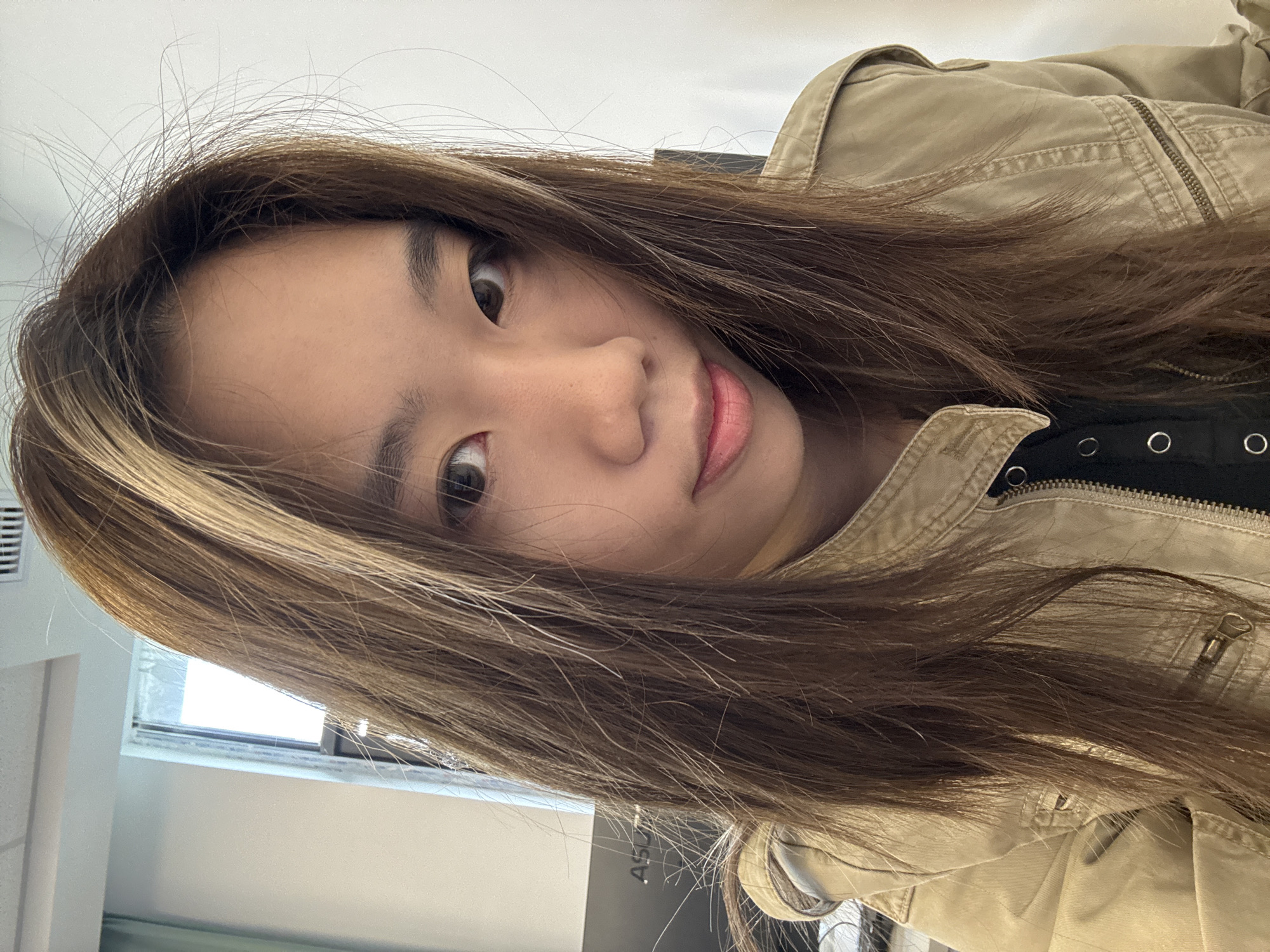A young Asian woman is posing for a photo. She has long brown hair with blonde highlights and is wearing a tan jacket over a black shirt. The background is a brightly lit indoor space with a window on the left and a white wall. There are no discernible features to confirm the location in Nan'an District, China.