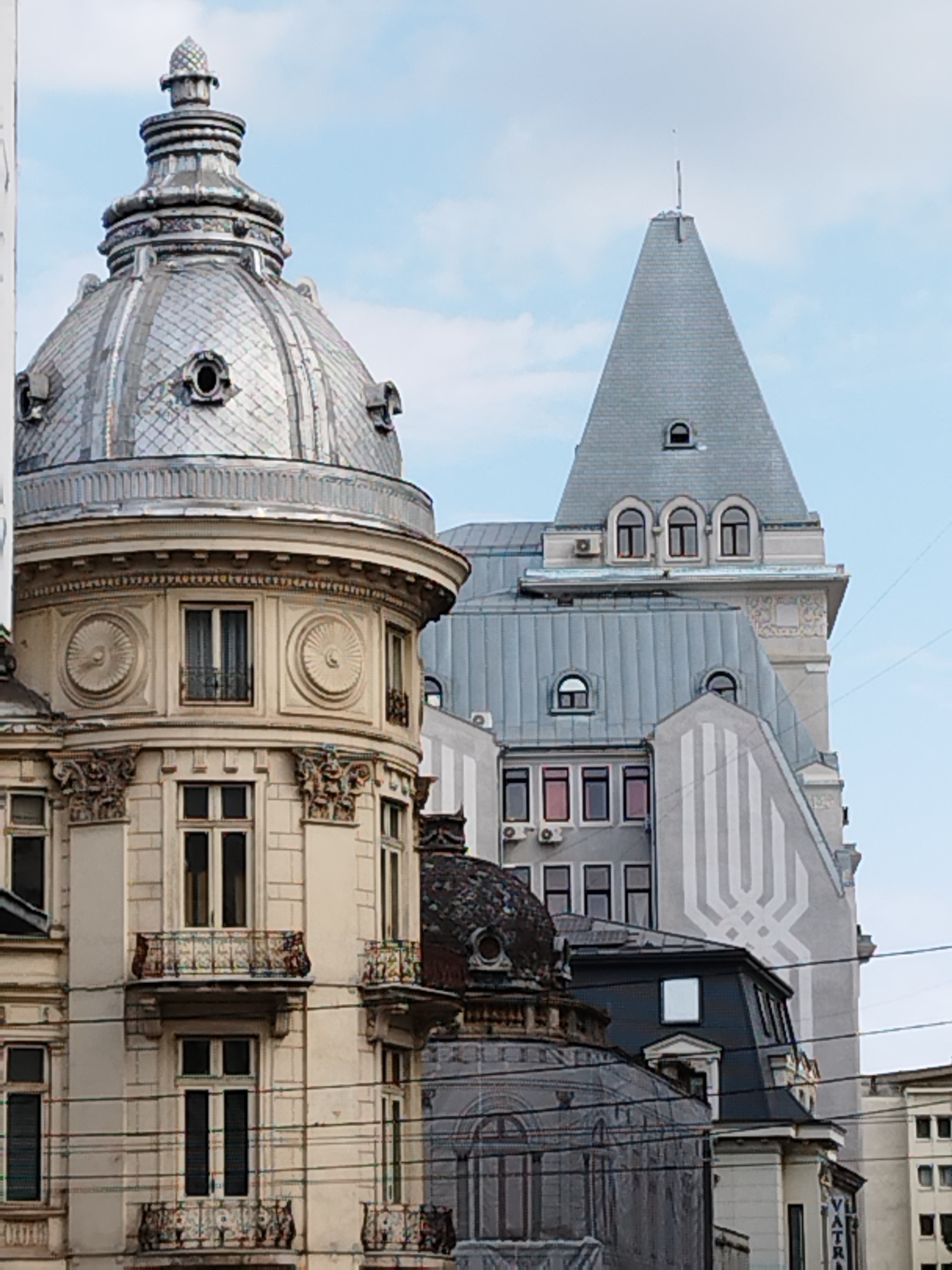 The image captures a close-up view of ornate architecture in Bucharest, Romania. In the foreground, a building with a large, silver, domed roof is visible. The dome is decorated with horizontal ribbed sections and features several small, circular windows. Below the dome, the building has a cream-colored facade with multiple windows, some of which are adorned with wrought-iron balconies. Decorative circular motifs are present above some of the windows, and elaborate carved details are visible on the corners of the structure.
Behind the domed building, a taller, grey building with a steeply pitched, slate-covered roof rises into the frame. This building has a tower-like element with arched windows at the top. Further to the right, another building with a similar grey, pitched roof is visible, featuring decorative grey patterns on its facade. The sky above is partly cloudy, with a pale blue hue. Electrical wires are strung across the scene, indicating an urban environment. The overall impression is one of historical architectural richness within the city.