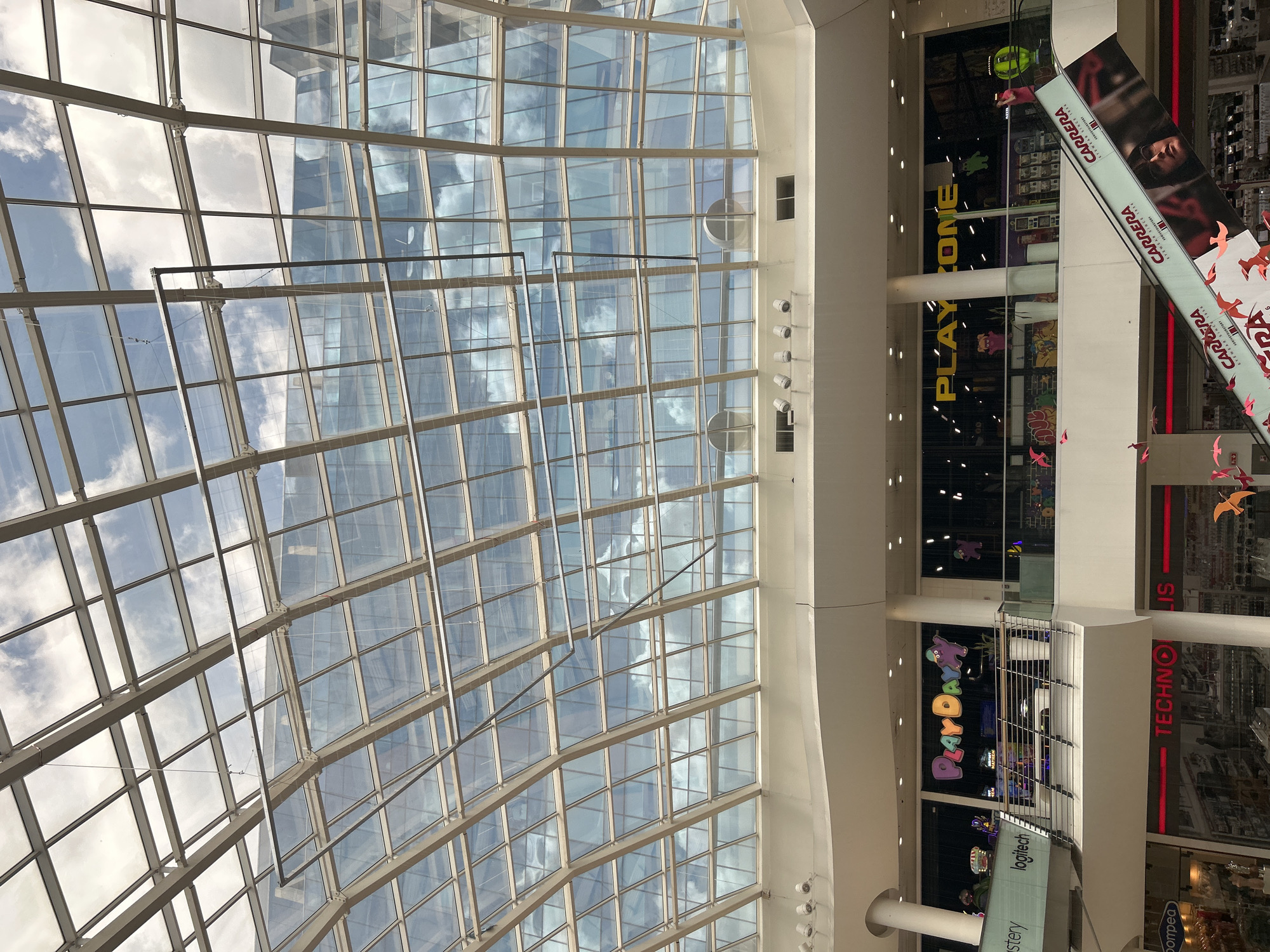 The image shows the interior of a shopping mall, with a distinctive glass roof structure composed of numerous rectangular panes reflecting the sky and clouds. The roof's support system is visible, consisting of a grid of metal beams and frames. Below the roof, the scene transitions to the interior levels of the mall. Various store signs are visible, including "PLAY ZONE", "PLAY DAY", "logitech", "TECHNO LIS", "pompea", and "CARRERA", each indicating different retail establishments. The mall's architecture includes white pillars and railings, suggesting a modern design. No people are clearly visible in the scene. The environment appears well-lit and clean.