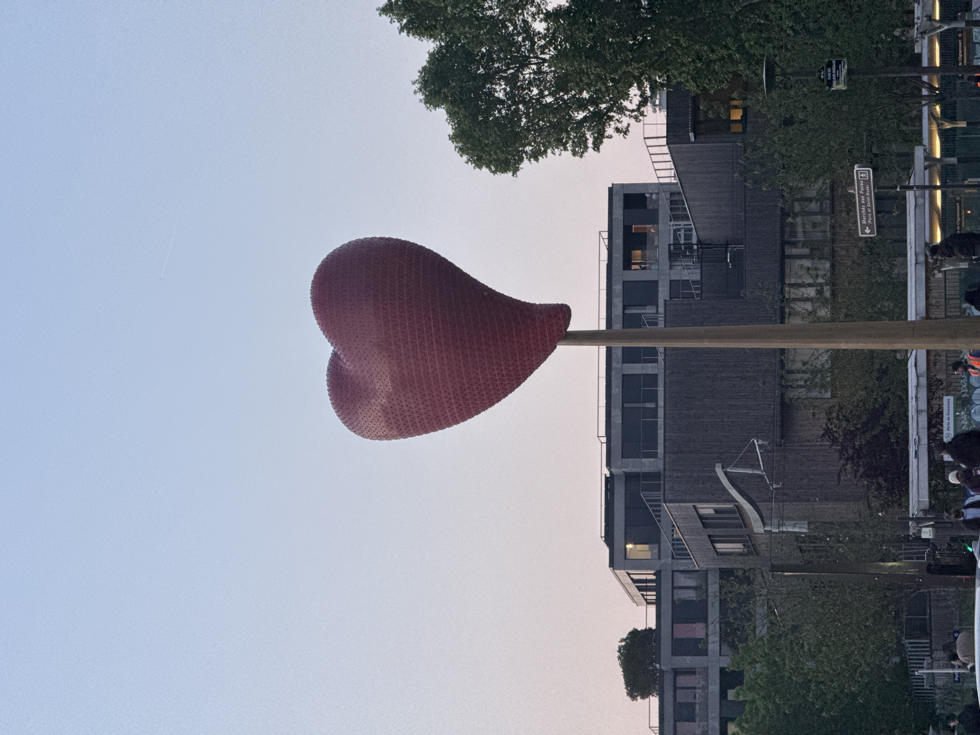 The image depicts a large, red, heart-shaped sculpture elevated on a pole against a backdrop of a pale sky and urban buildings in Paris, France. A street sign indicates "Marchés aux Puces - Porte de St-Ouen," a flea market location. Adjacent buildings appear modern, with visible windows and balconies. Trees are visible, partially obscuring some of the architectural details. A few people are present in the lower right, suggesting an urban environment.