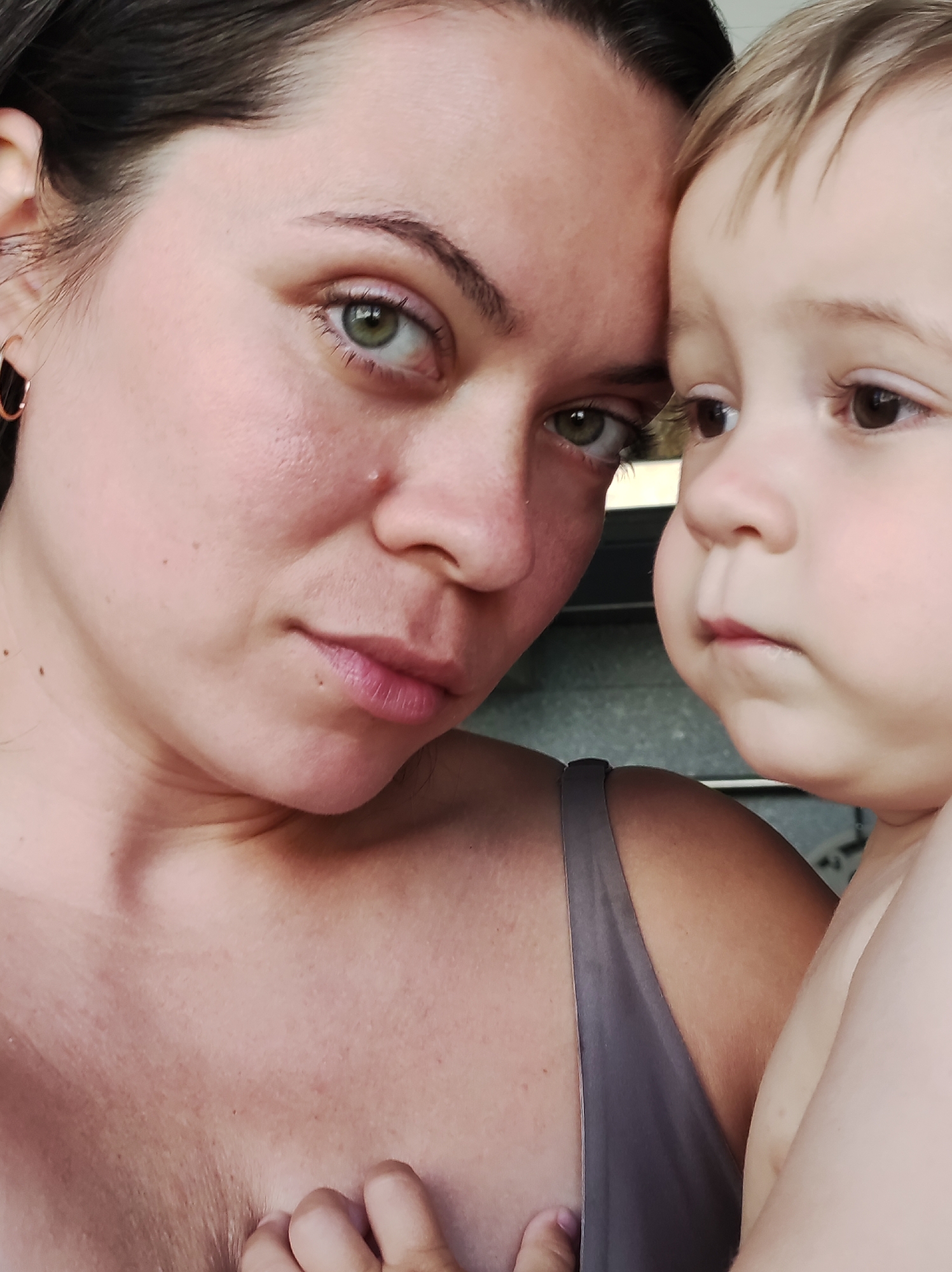 A close-up shot shows a woman and a young child in an indoor setting. The woman, with dark hair and green eyes, gazes directly at the viewer with a serious expression. She is wearing a gray top with thin straps. The child, who has light brown hair and brown eyes, is positioned beside her, looking off to the right with a similar expression of concentration. The background is blurry, but the setting appears to be simple. The composition is focused on the faces of the woman and child.