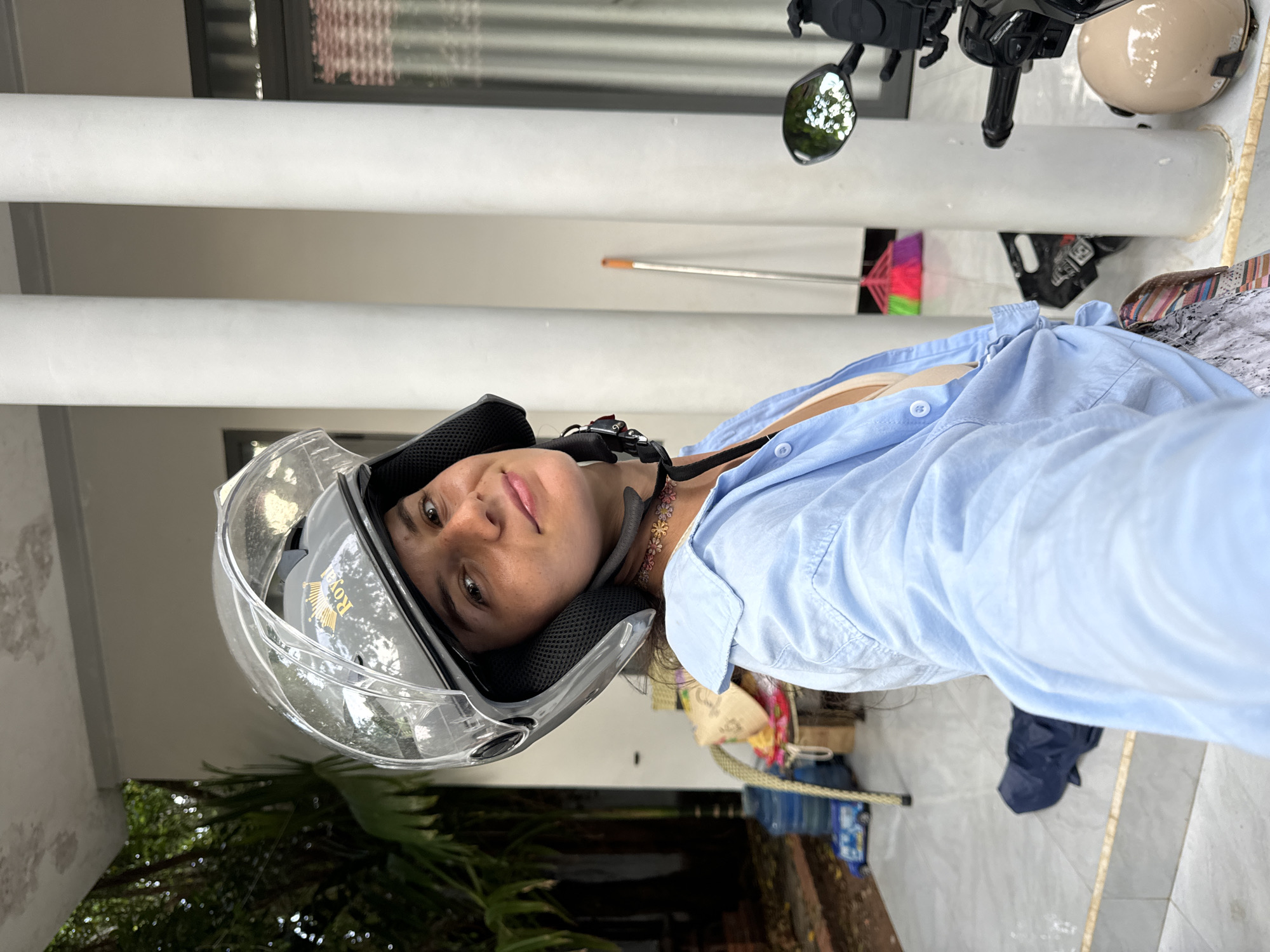 A woman wearing a light blue, open collared shirt and a tan bra, with a patterned skirt, is posing for a selfie. She has a grey motorcycle helmet on, with the clear visor up. Her hair is dark brown and pulled back. A delicate necklace with small floral charms is visible around her neck. She is looking directly at the camera with a slight smile.
The background suggests an outdoor covered area, possibly a porch or carport, in Mũi Né, Vietnam. White cylindrical pillars are prominent. To the left, there are large potted plants with lush green foliage, and a large blue water cooler. Various objects are scattered on the floor, including a colorful woven bag and what appears to be a child's toy truck. A broom with multi-colored bristles stands near the pillars. To the right, part of a motorcycle is visible, including the handlebars and a side mirror reflecting white lights. Another beige helmet is resting on a surface in the lower right corner. The ground is tiled.