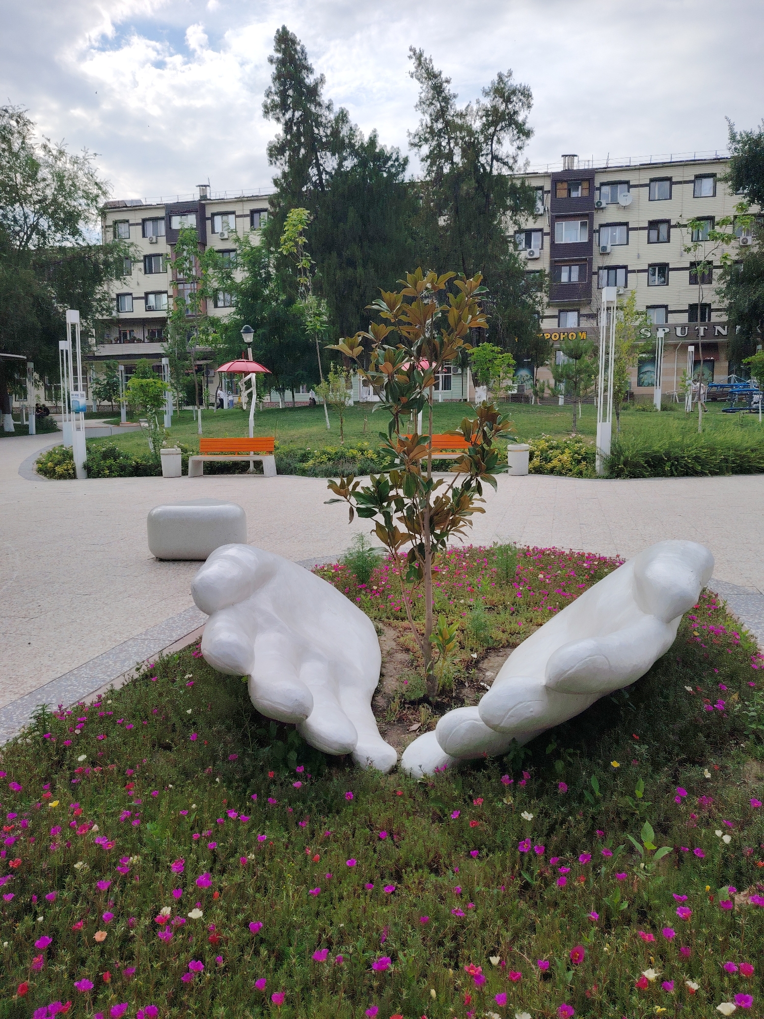 The image shows a park area in Shymkent, Kazakhstan, featuring a large white sculpture of two cupped hands in the foreground. The hands are placed within a flowerbed filled with small, colorful flowers in shades of pink, purple, and white. Behind the sculpture, a paved walkway curves through the park. In the midground, there is a small tree, a white cylindrical bench, and an orange bench. Further in the background, a multi-story building with multiple windows and air conditioning units stands behind a line of trees. The sky is partly cloudy. The building appears to have signage that includes Cyrillic text, and one sign reads "SPUTN".
