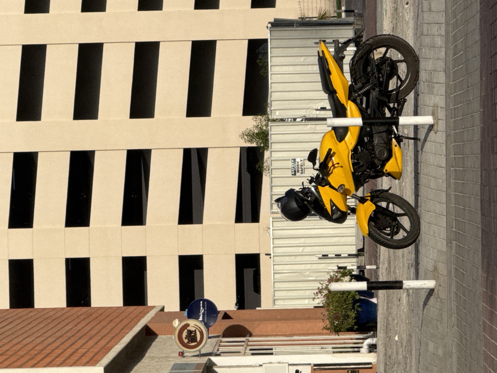 The image depicts a yellow motorcycle parked on the side of a street next to a building with multiple dark windows. A black helmet is attached to the motorcycle. The motorcycle is parked near a barrier with black and white stripes. The background shows the urban environment, likely Dubai, United Arab Emirates, based on architectural styles. The image is rotated ninety degrees, so the elements appear on their sides.