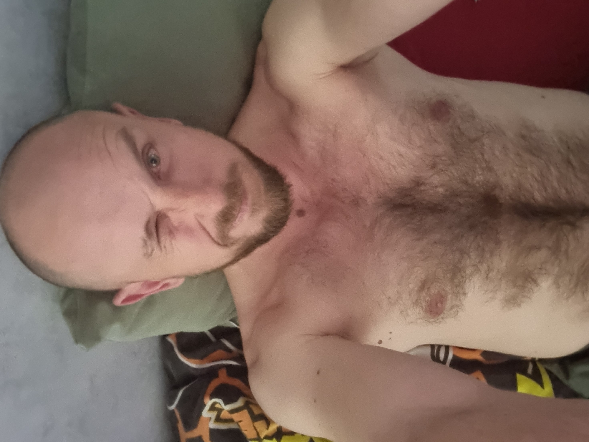 A shirtless man with a bald head, beard, and light skin is lying down. He appears to be on a bed with a green pillow and a colorful patterned blanket. The man is looking at the camera with one eye squinted. There is chest hair visible. The location is unknown and there is no indication of being in Sochi, Russia.