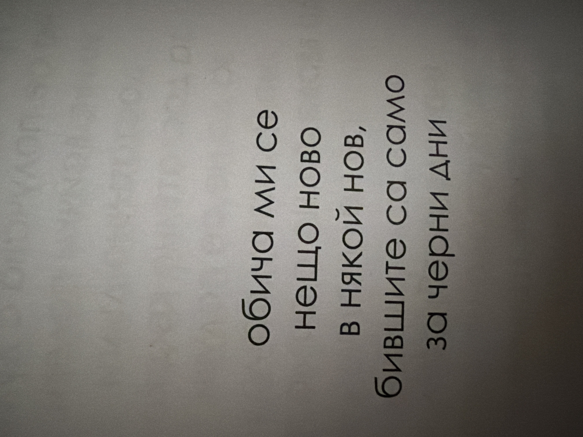 The image shows a close-up, angled view of text printed on a light-colored, textured surface, likely paper. The text is in Cyrillic script and appears to be a quote or poem. The words are arranged in five lines. The lighting is uneven, creating shadows and highlighting the texture of the surface. The overall impression is that this is a photograph of a printed page. There are no discernible elements that would allow for the identification of a specific location in Pazardzhik, Bulgaria, or any details about people, actions, or interactions.