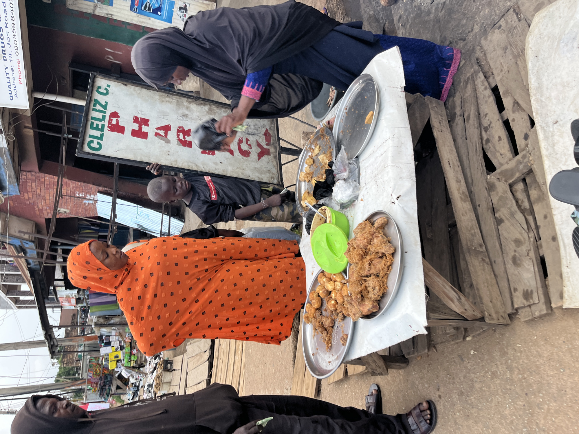 This image captures a street scene in Bukuru, Nigeria, where a woman in an orange headscarf and patterned dress stands beside a wooden stall laden with plates of fried food. In front of her, a young boy in a dark t-shirt and camouflage pants looks down, while a woman in a black hijab and dark blue skirt is seen on the right, holding a black plastic bag and interacting with the boy. A white sign with green and red lettering, reading "CLELIZ C. PHAR...", is propped up behind them, suggesting the proximity of a pharmacy. The stall is covered with a white cloth, and on it sit two metal plates filled with what appears to be fried chicken and possibly other fried items. A green plastic bowl is also visible. The background shows a busy marketplace with various stalls and structures. The ground is dirt, and the overall atmosphere suggests a vibrant, outdoor commercial setting.