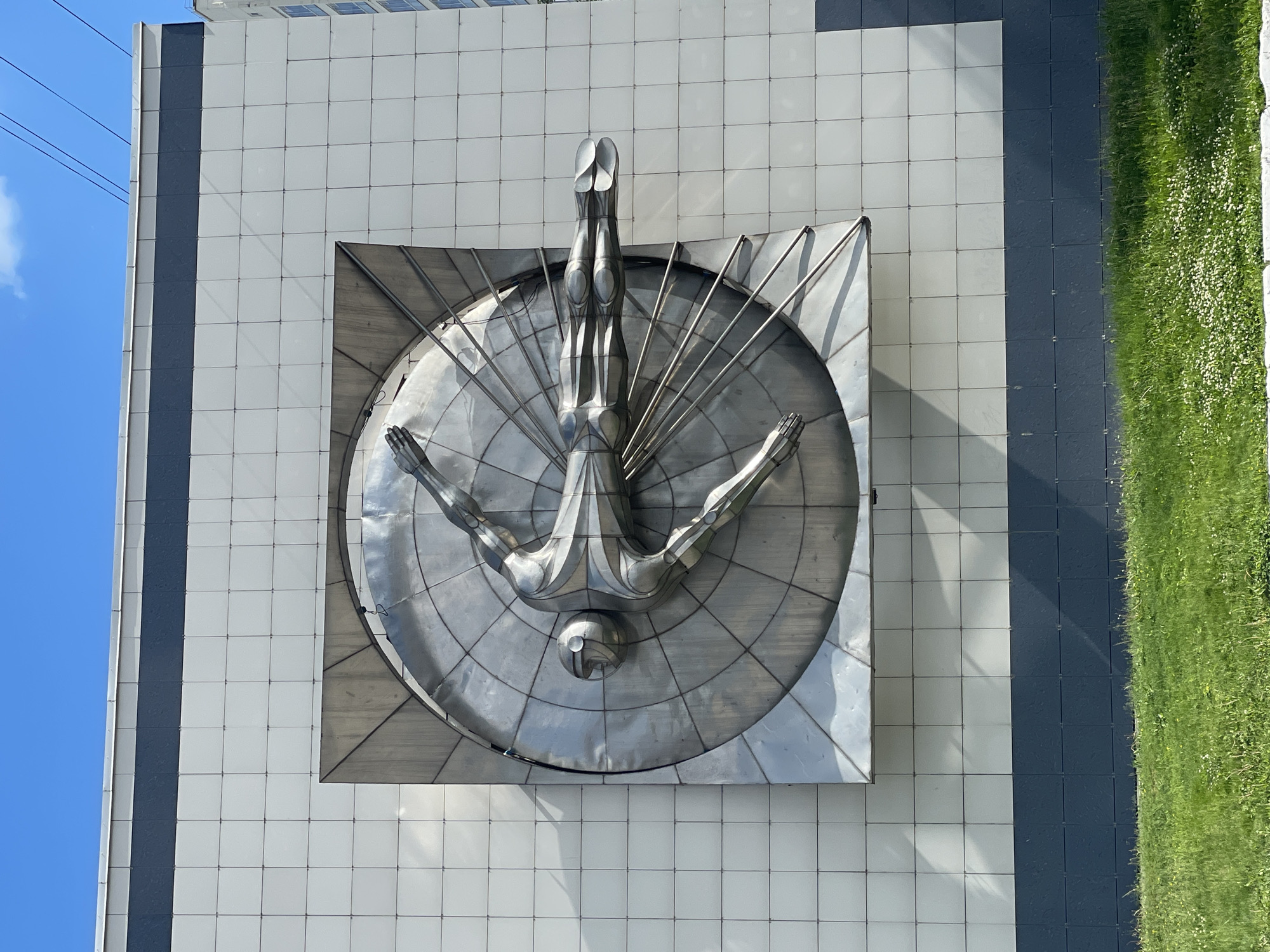 The image depicts a large, metallic sculpture of a human figure in motion, mounted on the facade of a modern building. The sculpture is silver and appears to be made of polished metal, with intricate details in the musculature and pose of the figure. The figure is positioned horizontally, with arms outstretched and legs extended, as if flying or running at high speed. The sculpture is set against a circular, radial background that further emphasizes the sense of speed and movement. The building's exterior is clad in light-colored square tiles, with a dark band running horizontally across the lower section. In front of the building, there is a strip of green grass with scattered white flowers. The sky above is clear and blue with some wispy clouds. This sculpture is located in the city of Perm, Russia, and is a prominent public artwork.