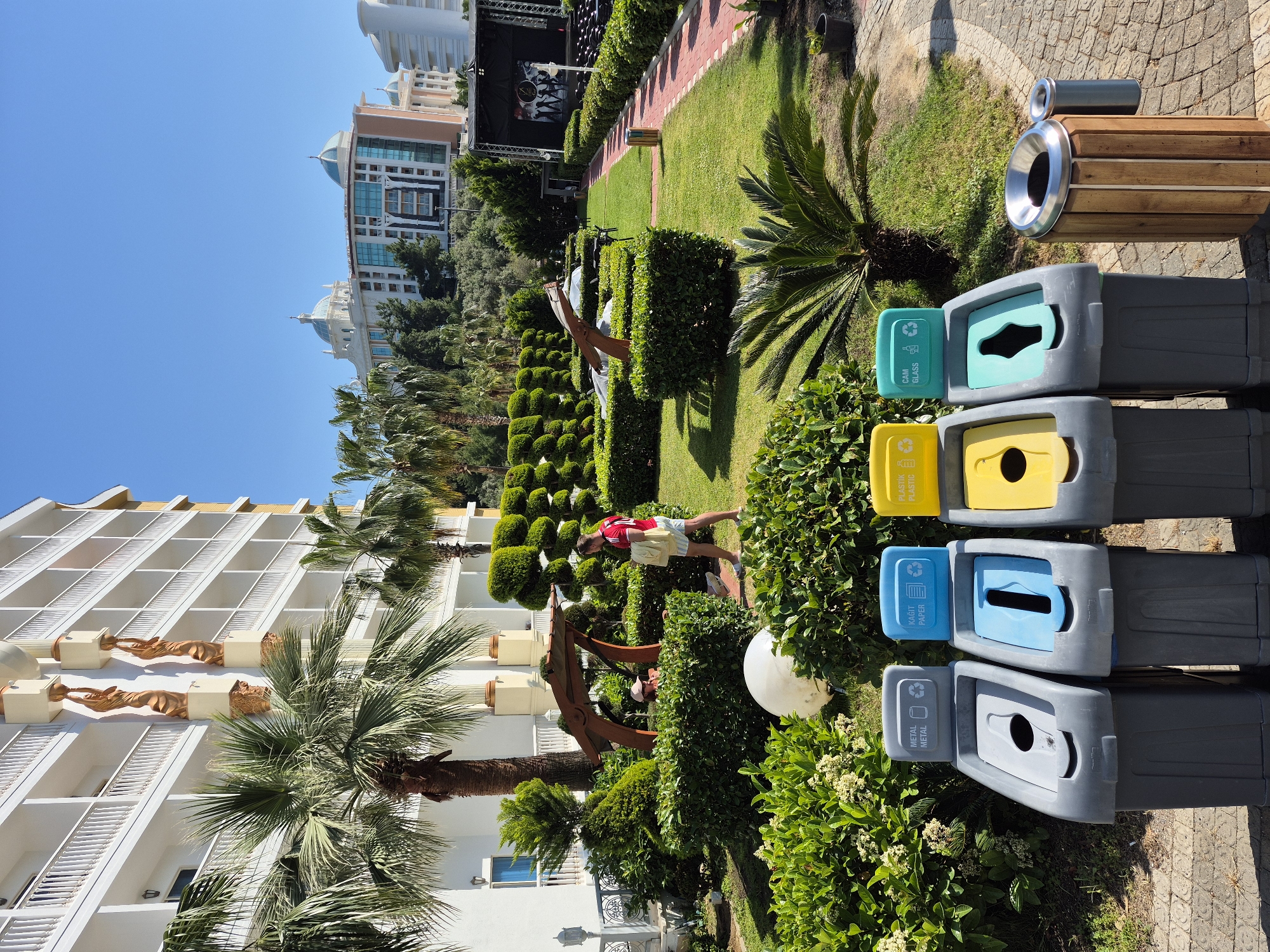 The image shows an outdoor scene in Alanya, Turkey, featuring a well-maintained garden and several recycling bins. The garden has manicured hedges, palm trees, and various plants. A man in a red shirt and light-colored shorts is standing near a swing or decorative structure within the garden.
In the foreground, there are recycling bins stacked vertically. These include bins for glass (cam), plastic (plastik), paper (kagit), and metal (metal). Next to them is a wooden bin for mixed waste.
In the background, there are large buildings, possibly hotels or resorts, under a clear blue sky. The overall scene suggests a tourist area in Alanya with attention to environmental practices through recycling.