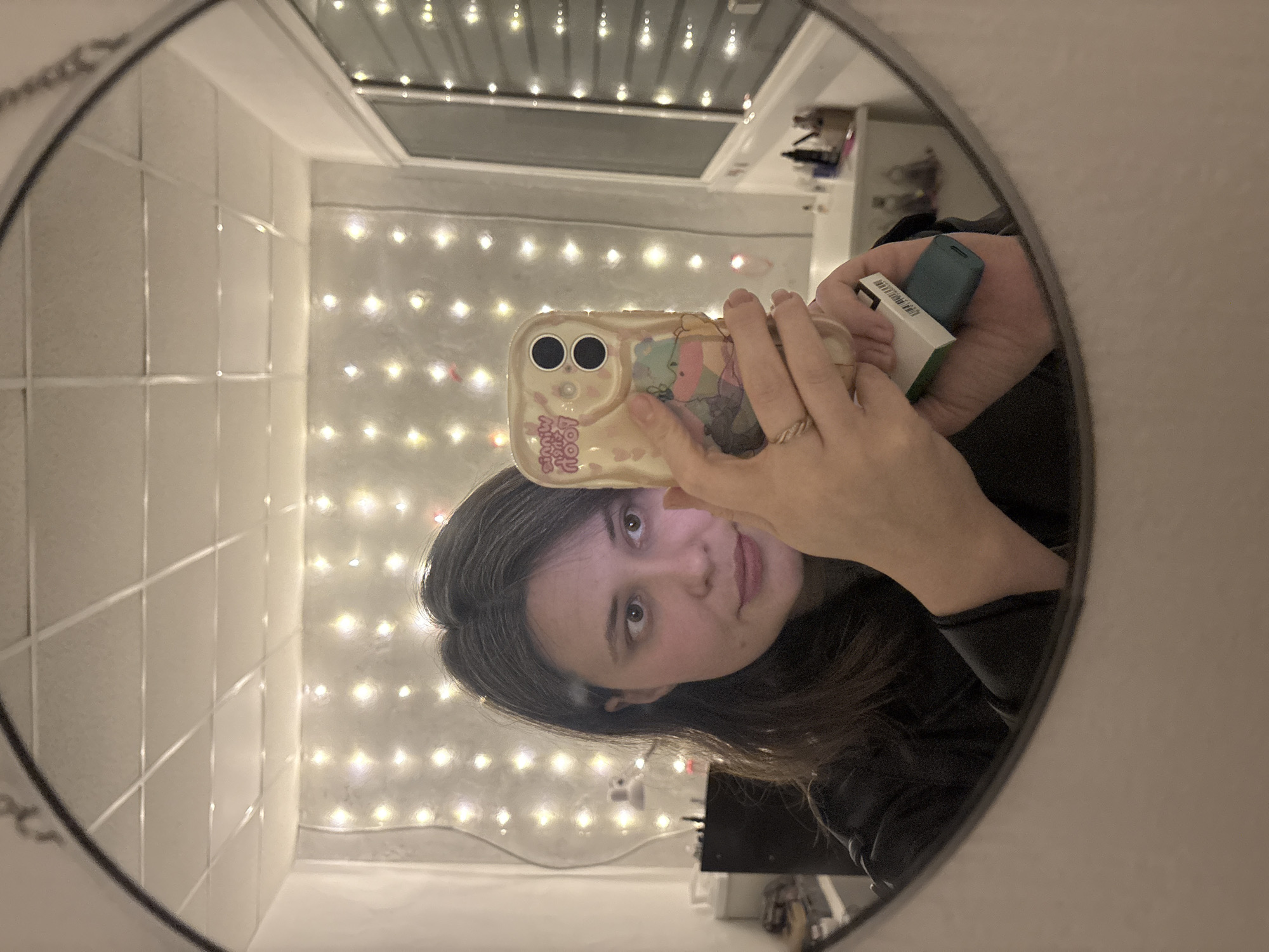 A female individual is depicted in a mirror selfie taken within a domestic interior in Chaikovsky, Russia. The individual, positioned centrally, has dark brown hair with visible gray strands, light-colored eyes, and is looking slightly to her right. She wears dark clothing, possibly a black jacket. Her left hand holds a light-colored smartphone with a decorated case featuring cartoon characters and partially visible text. A golden ring is present on her left ring finger. Her right hand, partially obscured by the phone, holds a small teal-colored electronic device, possibly a vaping device, partially enclosed in a white and green box.
The background, reflected in the mirror, shows a room illuminated by numerous string lights hung across a light-colored wall. The ceiling features a grid pattern, indicative of a drop ceiling with square panels. To the left, a dark piece of furniture and a white cylindrical object are visible. To the right, a window with vertical elements (blinds or grates) reflects the string lights. Below the window, a white vanity or counter holds various toiletries and bottles, with a white bathtub or sink basin visible on the far right.