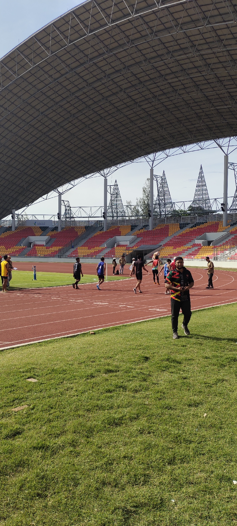 An outdoor, vertical, medium shot captures a view of a modern stadium in Banda Aceh, Indonesia, on a bright, sunny day. The stadium features a large, grey, arched roof supported by white pillars and intricate metal frameworks. Red and yellow tiered seating is visible beneath the roof, some of which appears empty. A red running track encircles a green field of grass.
On the track, several individuals are engaged in athletic activities. In the foreground, a man in a black shirt with colorful geometric patterns walks towards the camera. Behind him, a group of young people are seen on the track. Some are running, while others appear to be walking or observing. One individual in a black shirt with the number "11" on the back is running, and another in a blue shirt with a number "1" is also running. Further back, other participants and spectators are dispersed on the track and in the stands. A checkered flag is held by one of the individuals on the track. The environment appears to be a sporting event or training session.