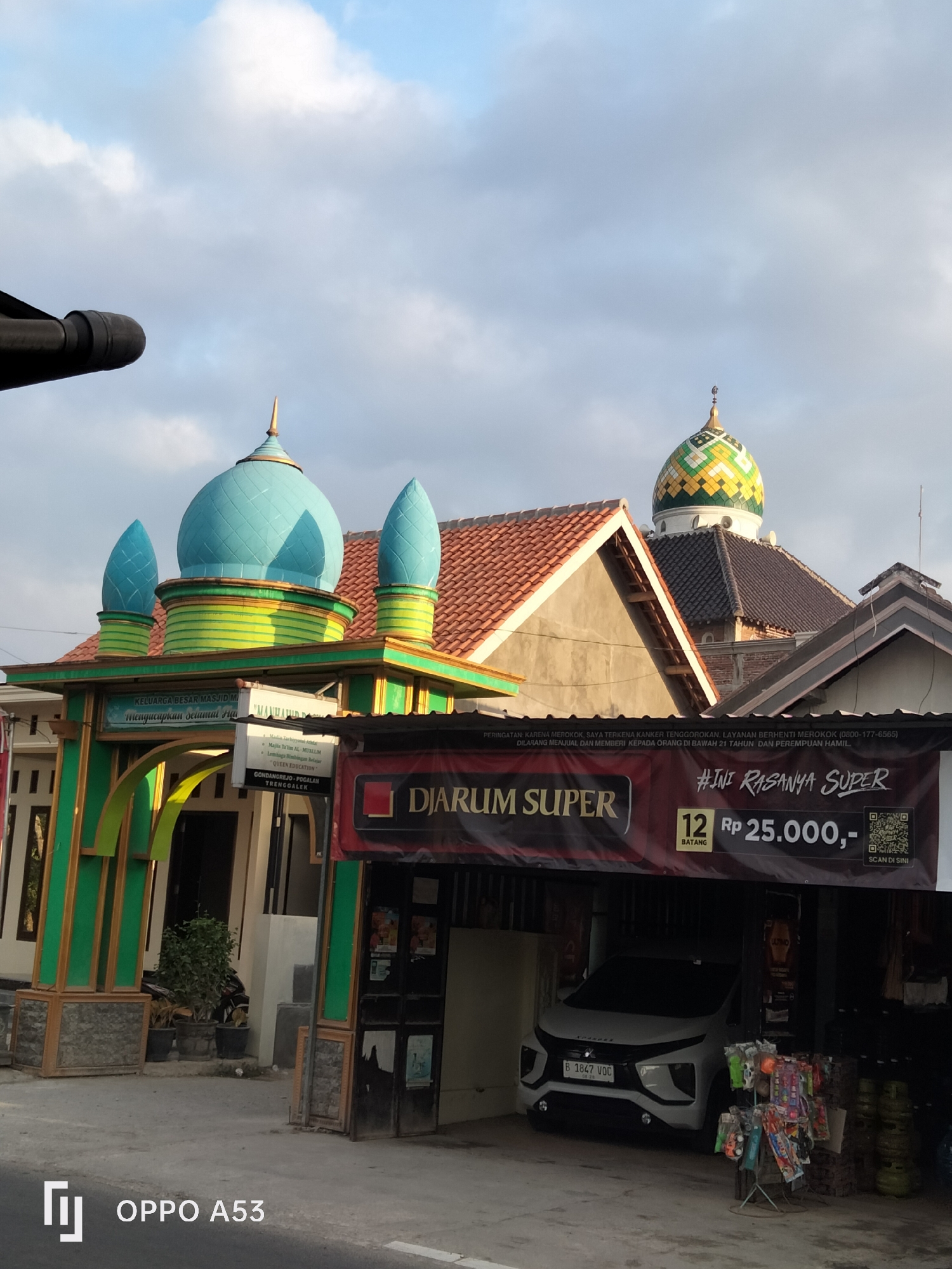The image shows an exterior view of what appears to be a mosque entrance and a storefront in Trenggalek, Indonesia. The mosque entrance is adorned with green and gold decorations and features two large blue domes and two smaller blue spires. A banner above the entrance reads "KELUARGA BESAR MASJID M..." and "Mengucapkan Selamat Ha...". Behind the mosque entrance, a larger dome with a mosaic of green, yellow, and white tiles is visible, topping a building with red-tiled roofs. To the right of the mosque, a storefront displays a large banner with the text "#INI RASANYA SUPER" and "12 BATANG Rp 25.000,-". Below this banner, a white car is parked inside the store, and a display rack with various colorful items is positioned in front of it. The environment appears to be a street scene under a partly cloudy sky. The lower right corner of the image includes the text "OPPO A53", indicating the device used for capture.