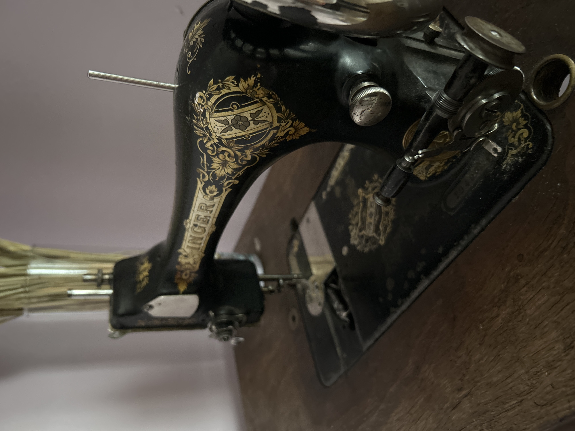 The image depicts an antique black Singer sewing machine with gold decorative accents. The machine sits on a dark wooden surface, likely a table or stand. The background is a blurred, light-colored wall. Details of the machine include the "Singer" name in gold lettering on the arm, and various knobs and mechanisms for sewing. The location is unknown, the prompt "city of Chaikovsky, Russia" is assumed unrelated.