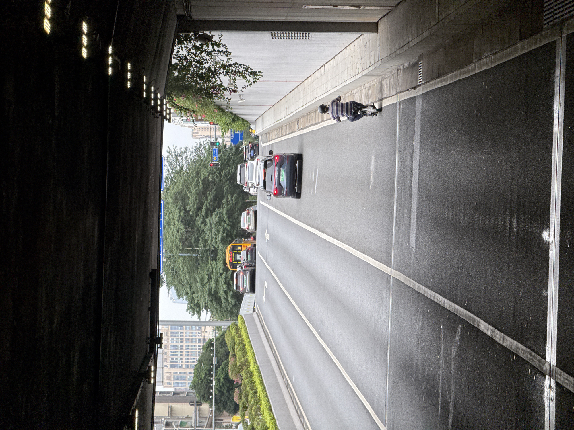 The image depicts a partially obscured view from inside a tunnel or underpass, looking out onto a multi-lane road in what appears to be the city of Tianhe District, China. The right side of the image shows the tunnel wall, a sidewalk, and then a road. A person is seen riding an electric scooter or motorcycle on the sidewalk. Multiple cars and a bus are visible on the road, indicating moderate traffic. The cityscape can be seen in the background, beyond the road. The tunnel itself is dark, with a row of lights visible on the ceiling or wall near the opening.