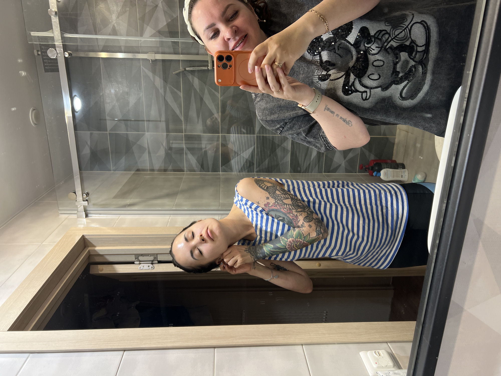 The image shows a mirror selfie taken in a bathroom. A woman is taking the photo. Her reflection shows a woman with tattoos on her arm, wearing a striped blue and white top. She appears to be putting on an earring. The bathroom contains gray patterned tiles behind the woman taking the selfie, a glass shower, a medicine cabinet and a light wood vanity. The image was taken in Noviy, Russia.