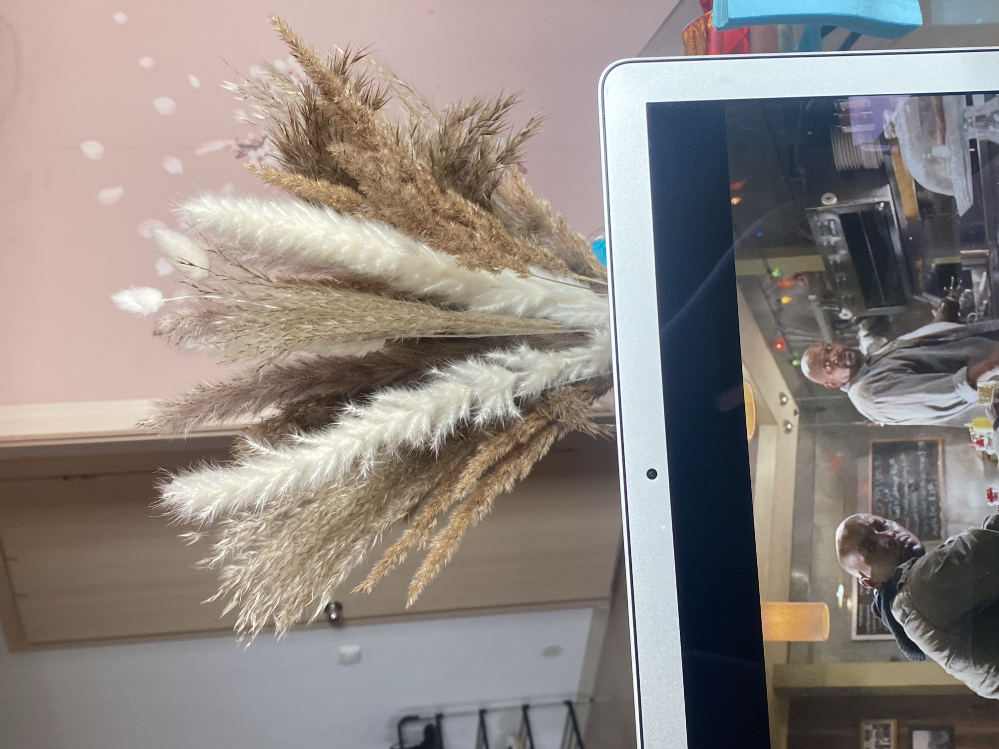 A vase containing beige and white feathery reeds stands on a surface, positioned near a computer screen displaying a film scene with actors. The scene appears to be indoors, possibly a bar or restaurant. The surrounding environment is blurry. The location in Yekaterinburg, Russia, is not discernible from the image.